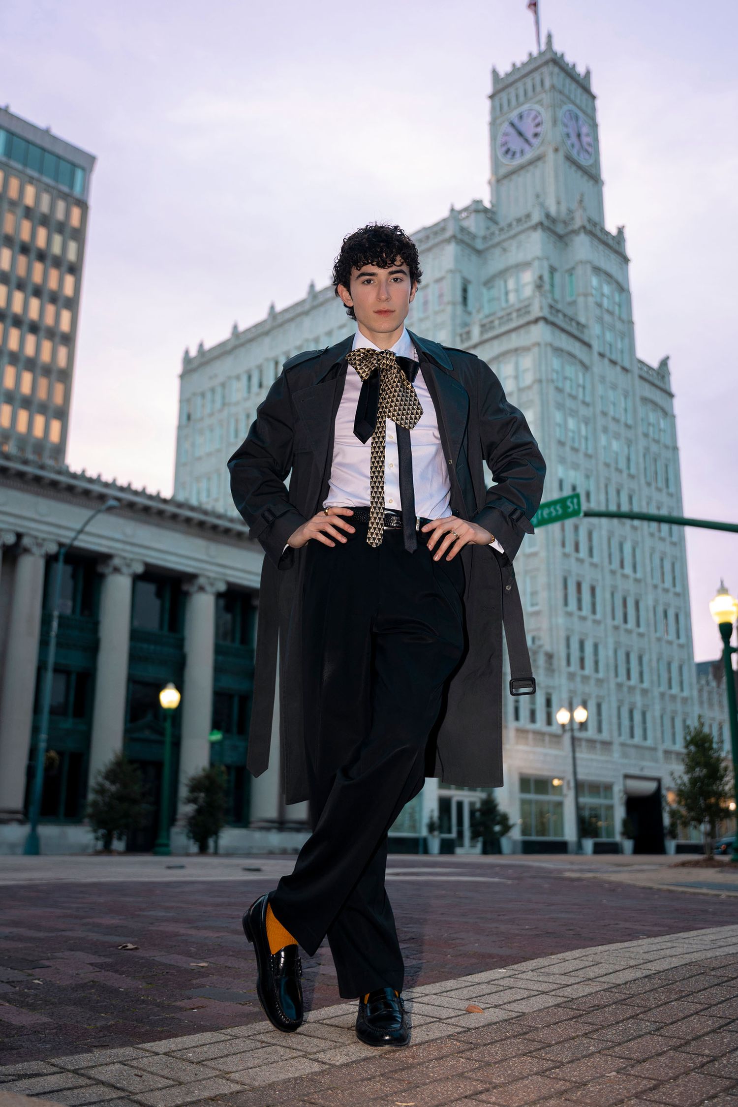 Young man in black trench coat, white shirt and gold patterned tie posing in urban plaza with clock tower.