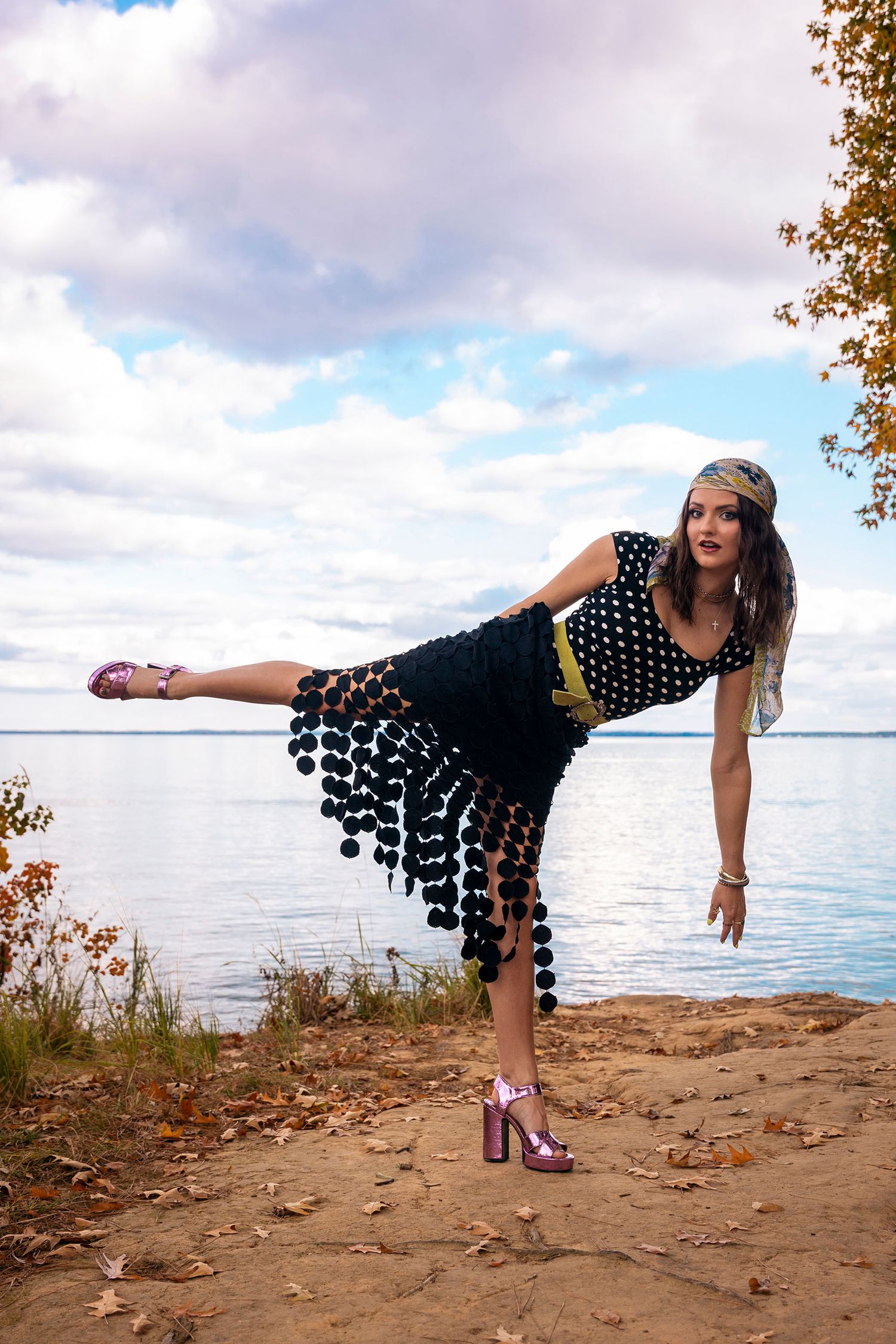 Woman in polka dot pom-pom dress balancing on one leg on sandy lakeside shore.