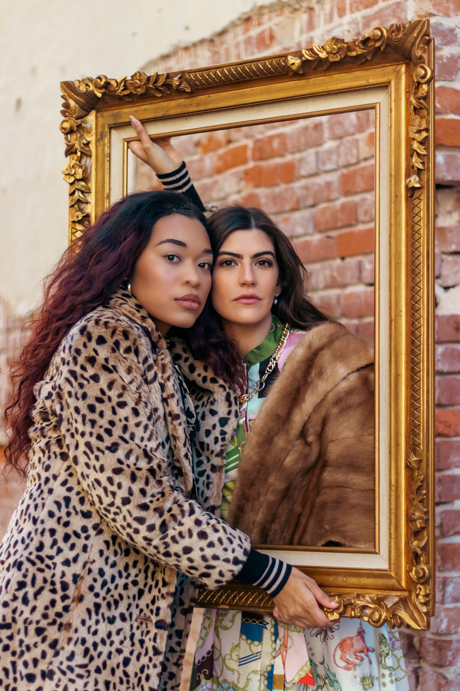 Two women in fur coats pose together holding a large ornate gold picture frame.