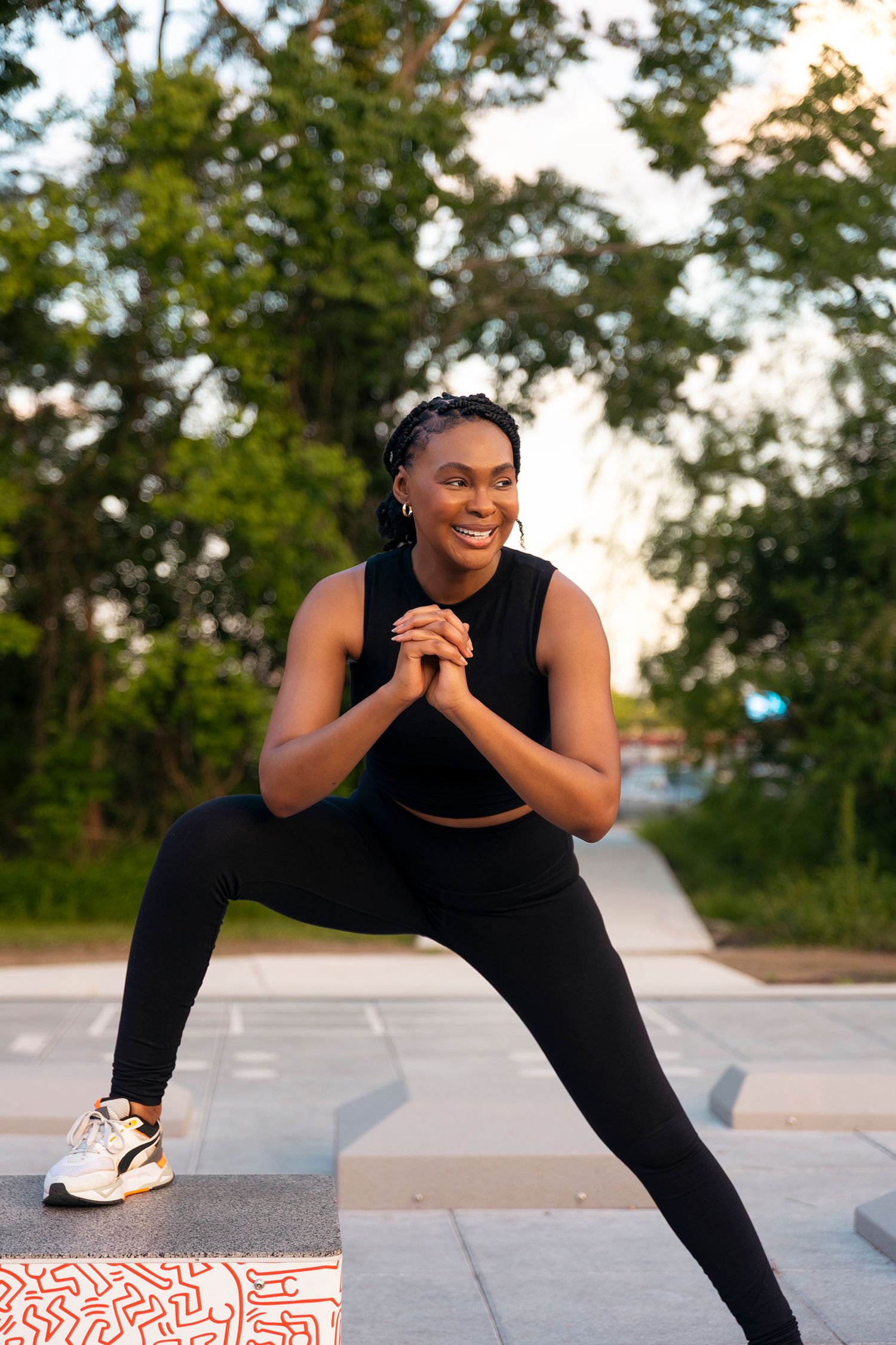 A smiling Black woman in black workout attire does a side lunge stretch in an outdoor park.