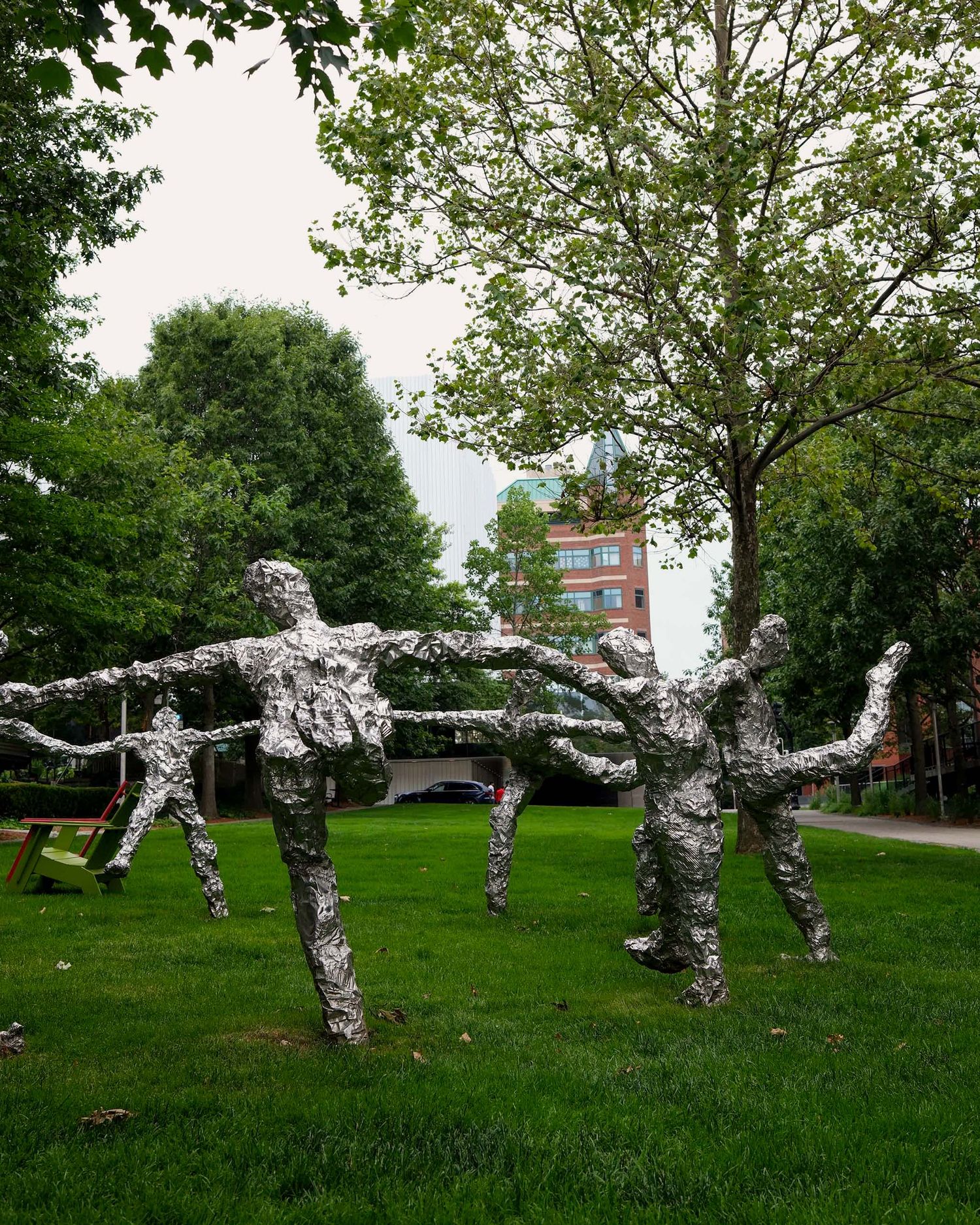 Silver metallic human figure sculptures holding hands on a green lawn in an urban park.