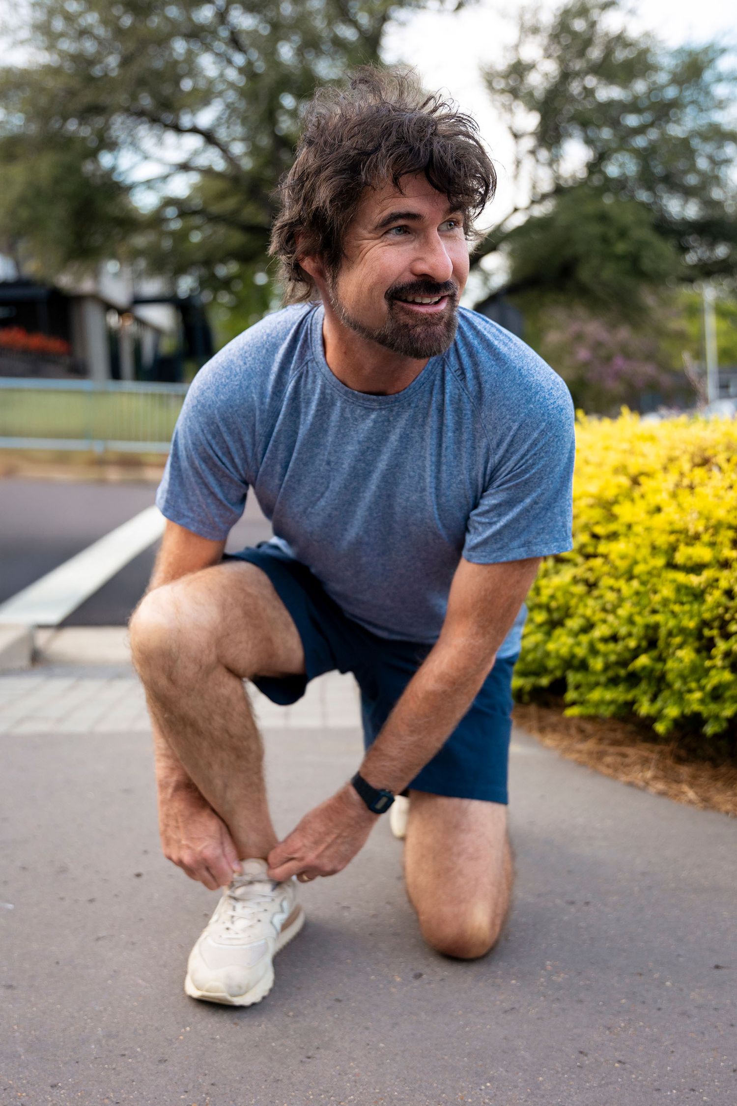 A smiling man in a blue t-shirt and navy shorts ties his white sneakers on a suburban street.