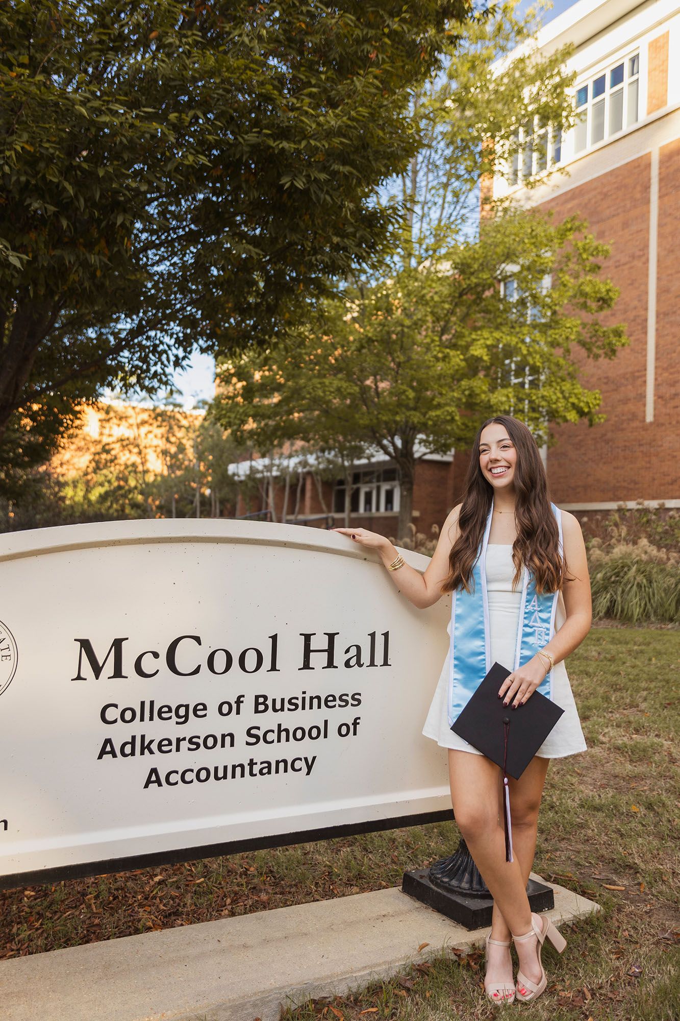 Happy graduate posing beside McCool Hall Adkerson School of Accountancy sign on campus.
