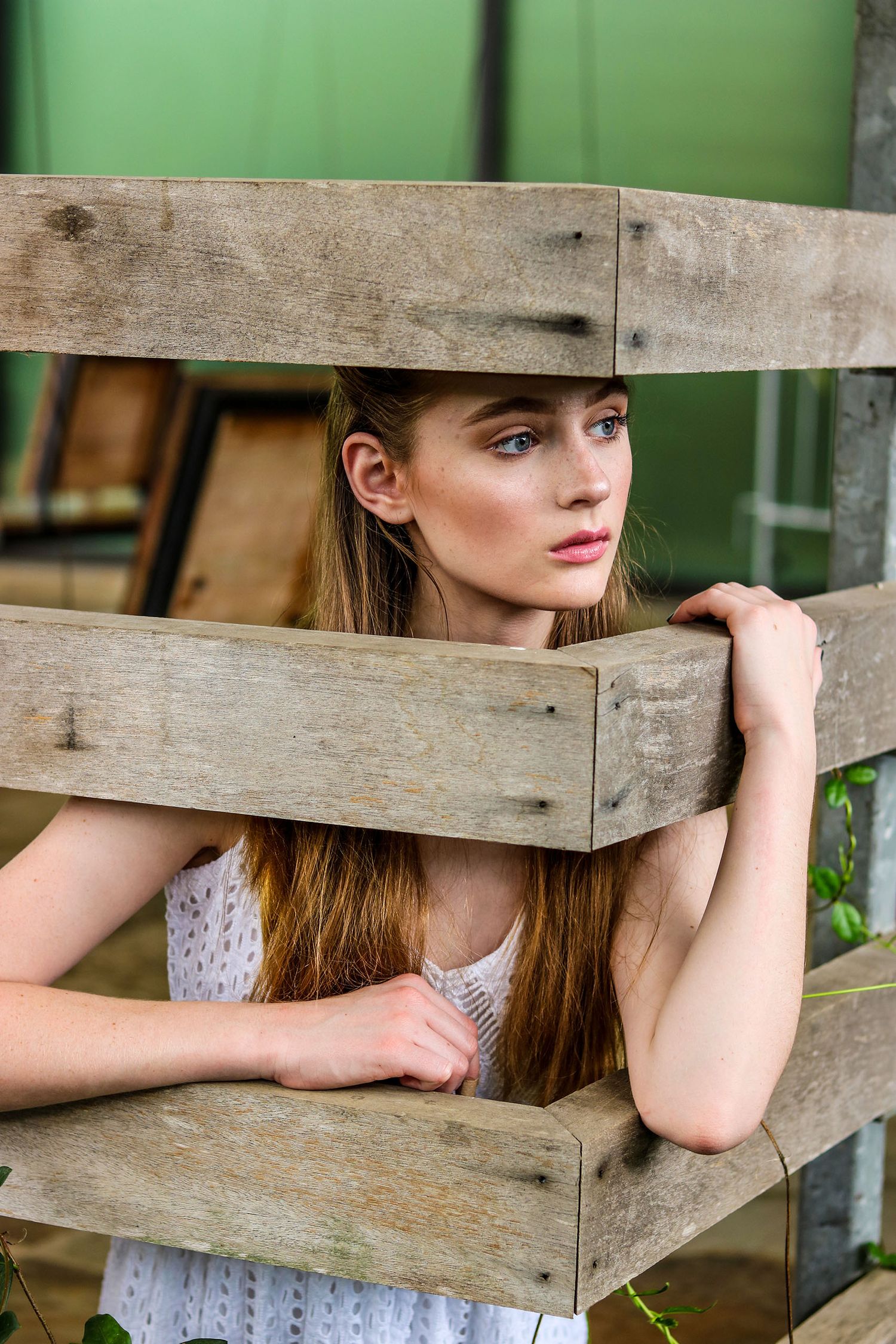 A young woman with long hair looks through gaps in a rustic wooden fence.