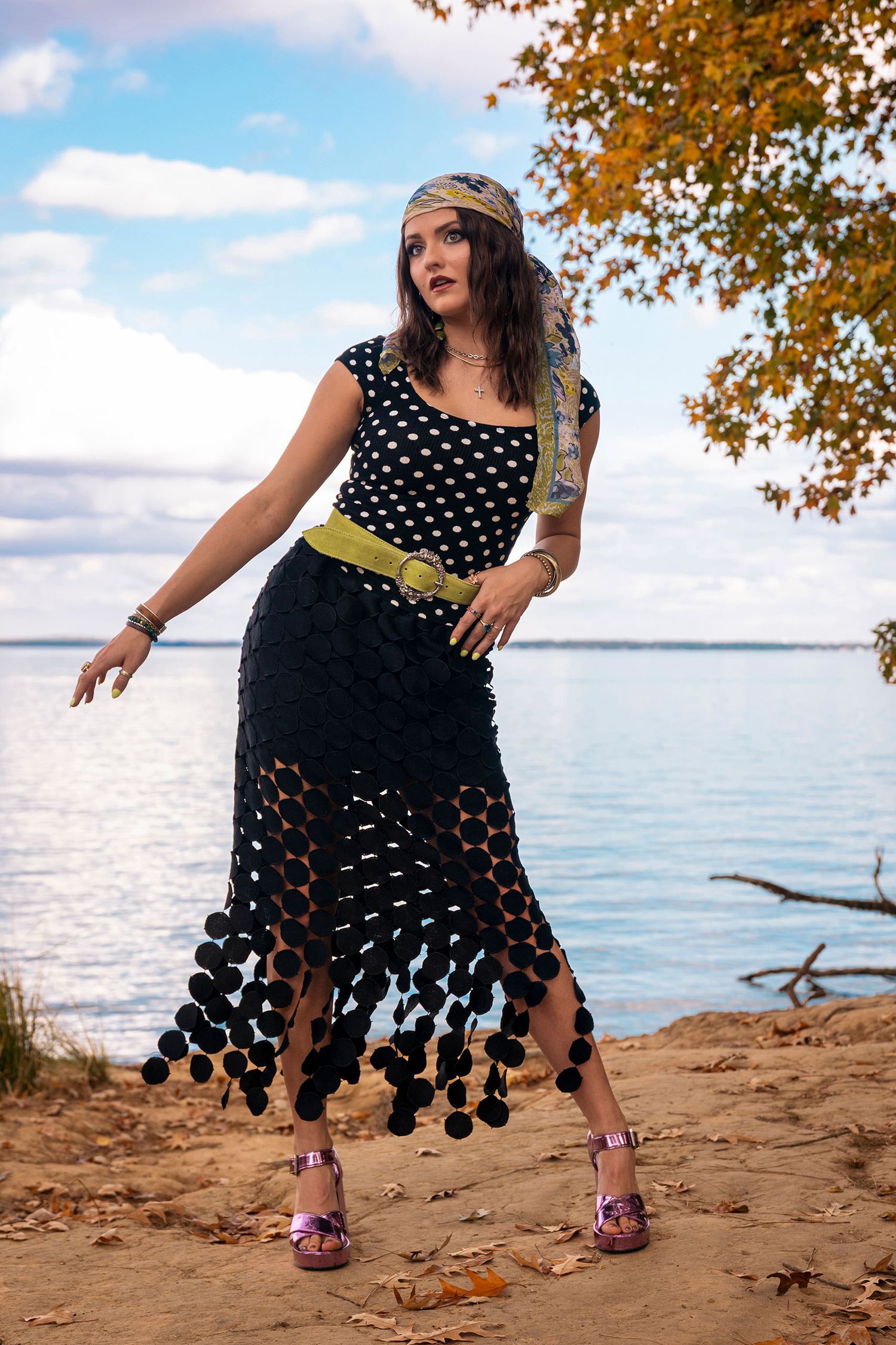Woman in polka dot dress with pom-pom hem and headscarf posing on sandy lakeside shore.