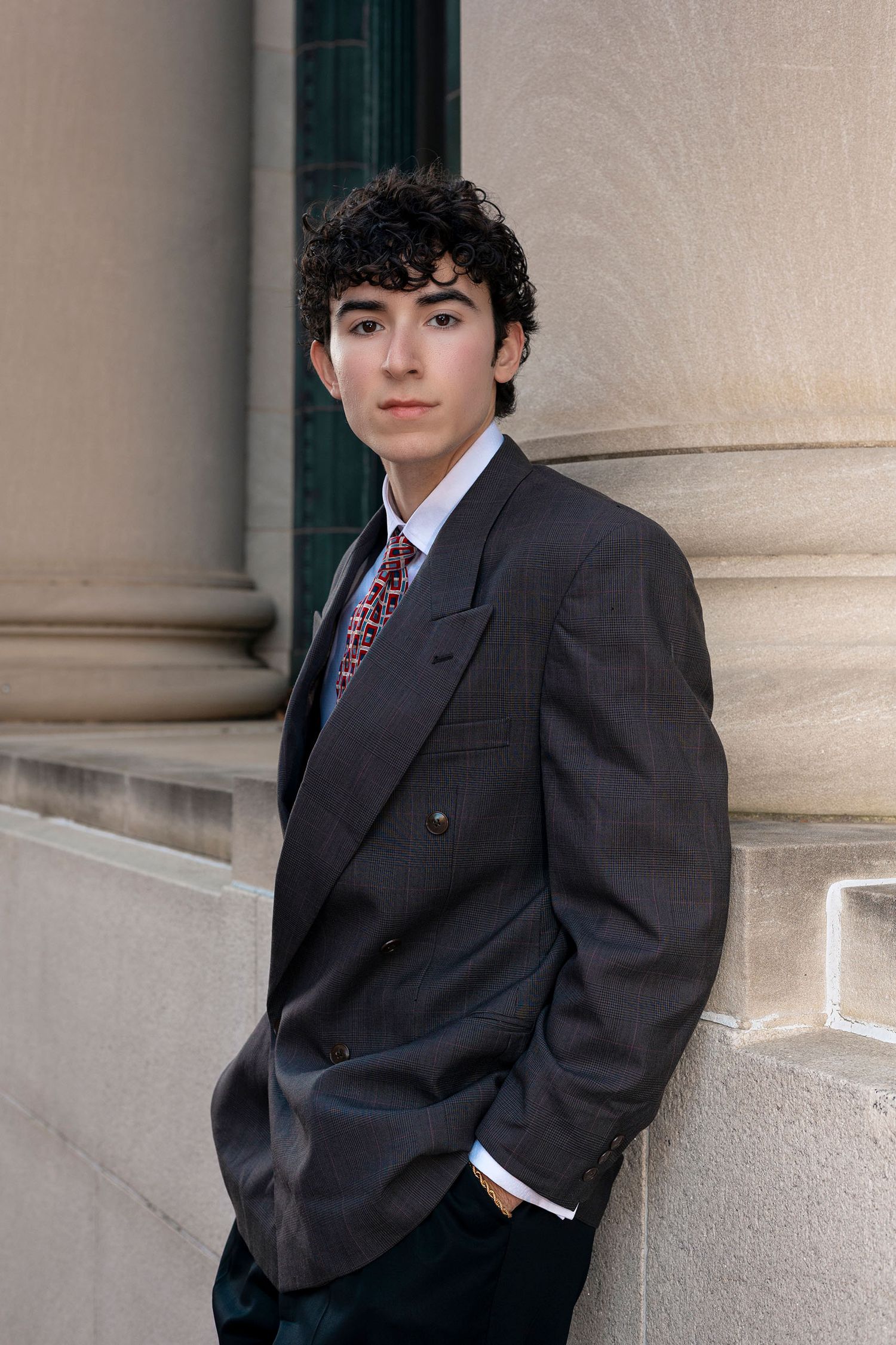 Young man in gray double-breasted suit and patterned tie leaning against stone column in classic portrait pose.