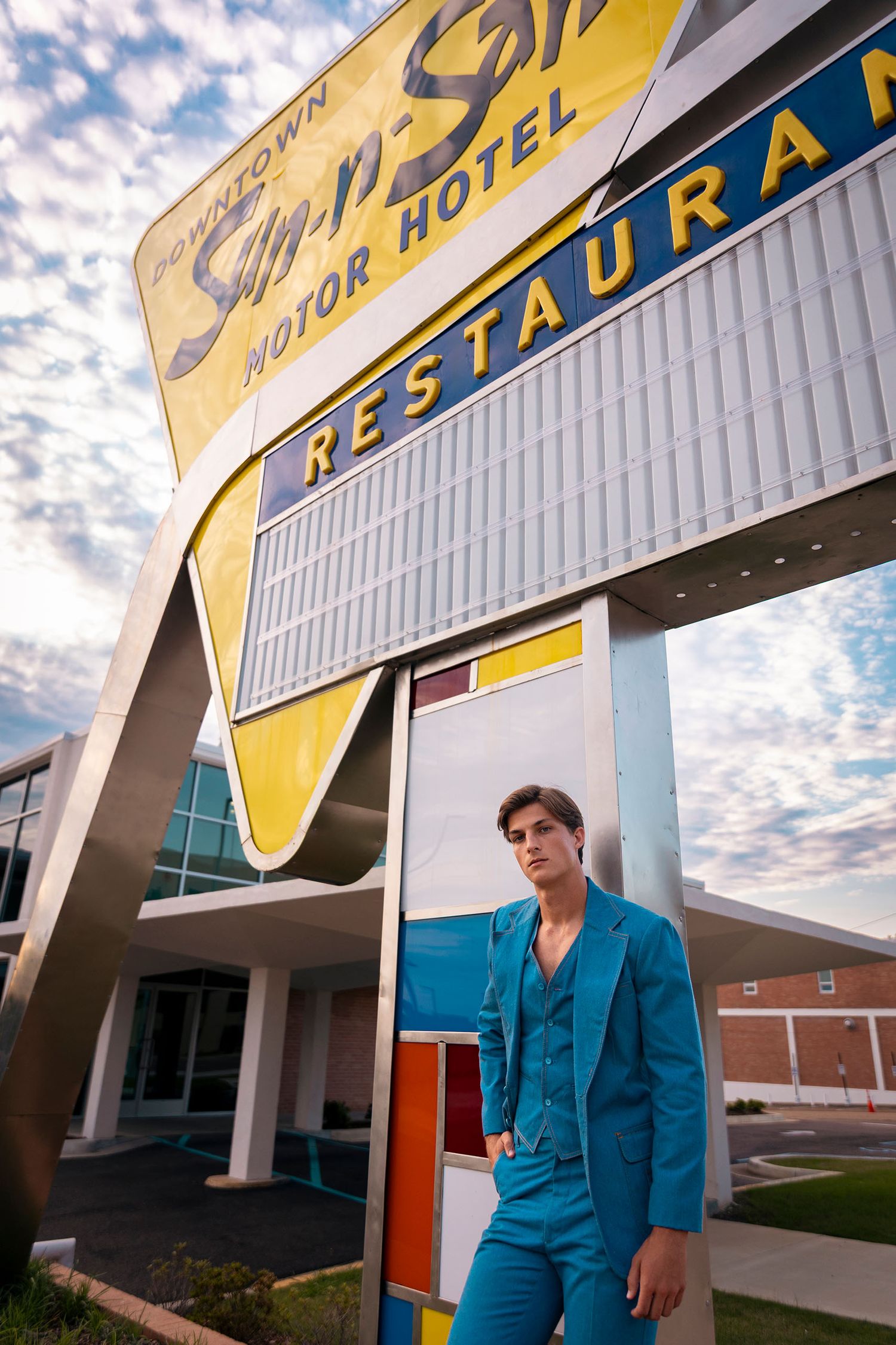Man in teal suit poses under vintage Downtown Motor Hotel Restaurant sign at dusk.