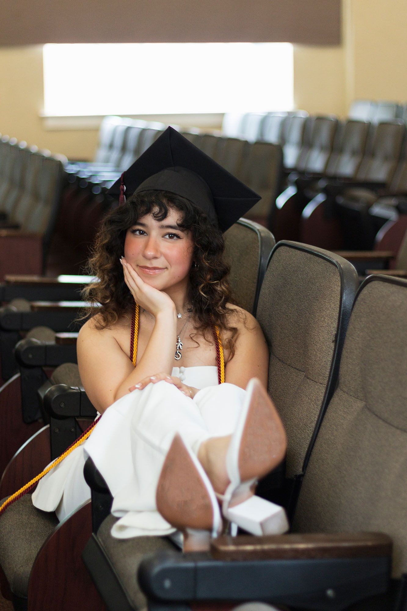 Female graduate in white dress sits in auditorium wearing graduation cap with honor cords.