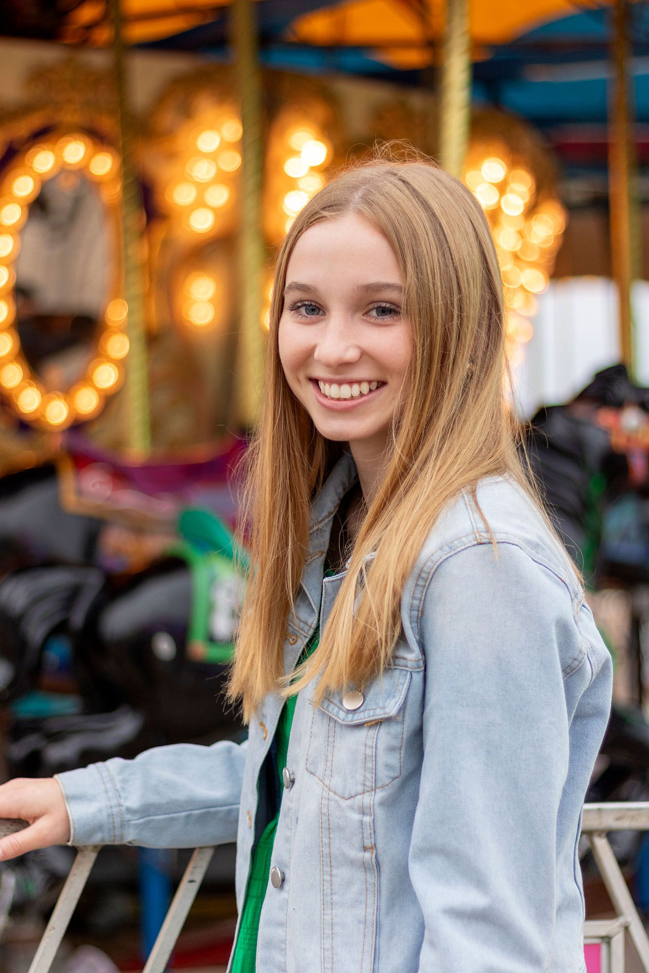 Smiling teenage girl in denim jacket at colorful carnival with carousel lights in background.