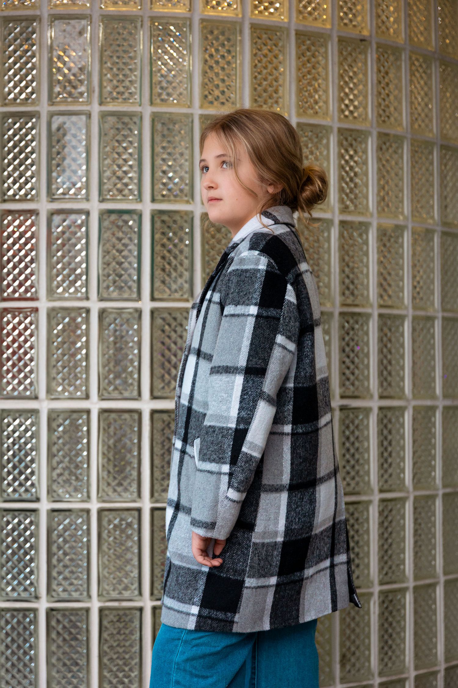 A young girl poses sideways in a black and white plaid oversized coat against a glass block wall.