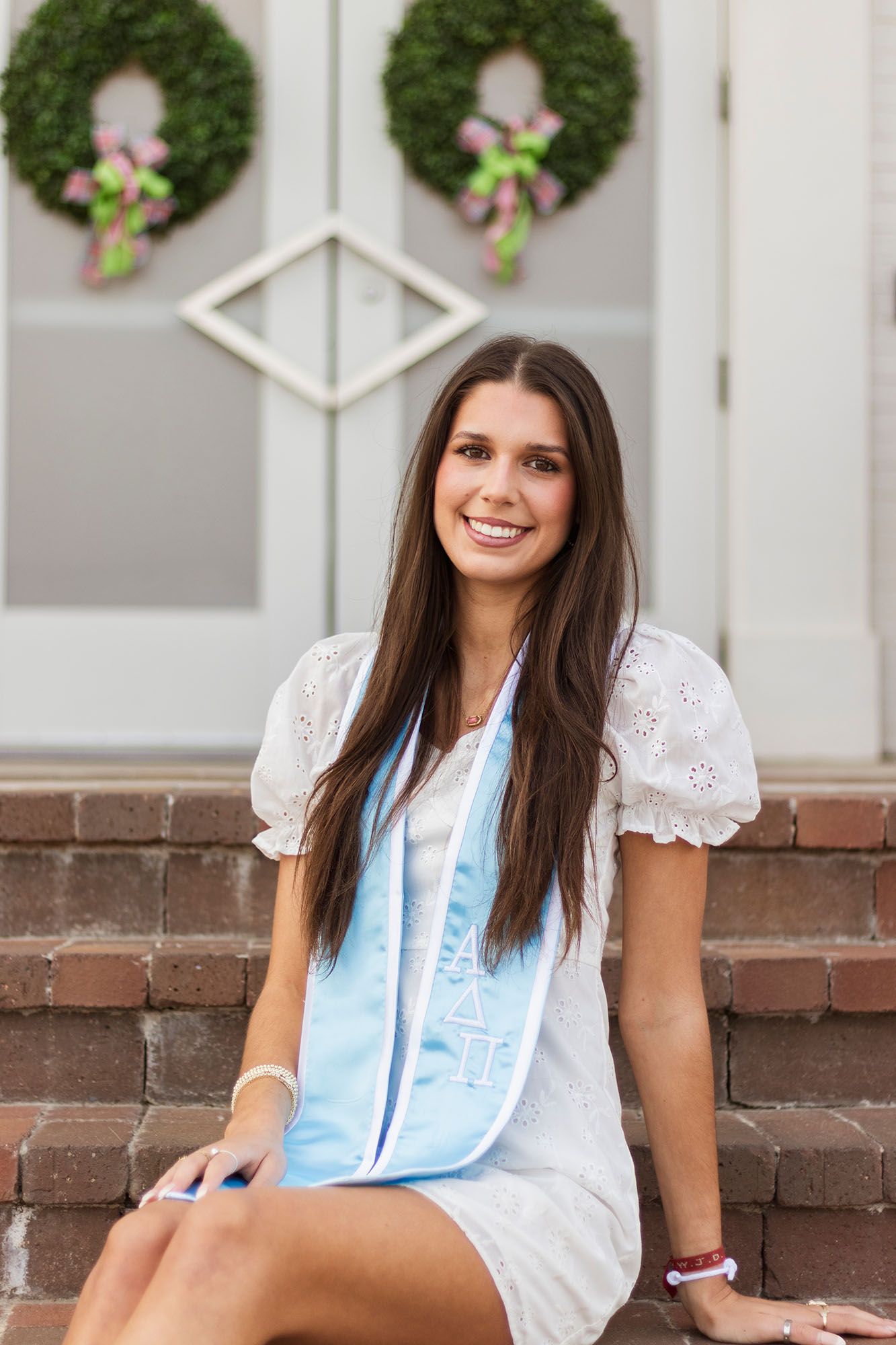 Brunette female graduate in white dress with light blue stole sits smiling on brick steps.