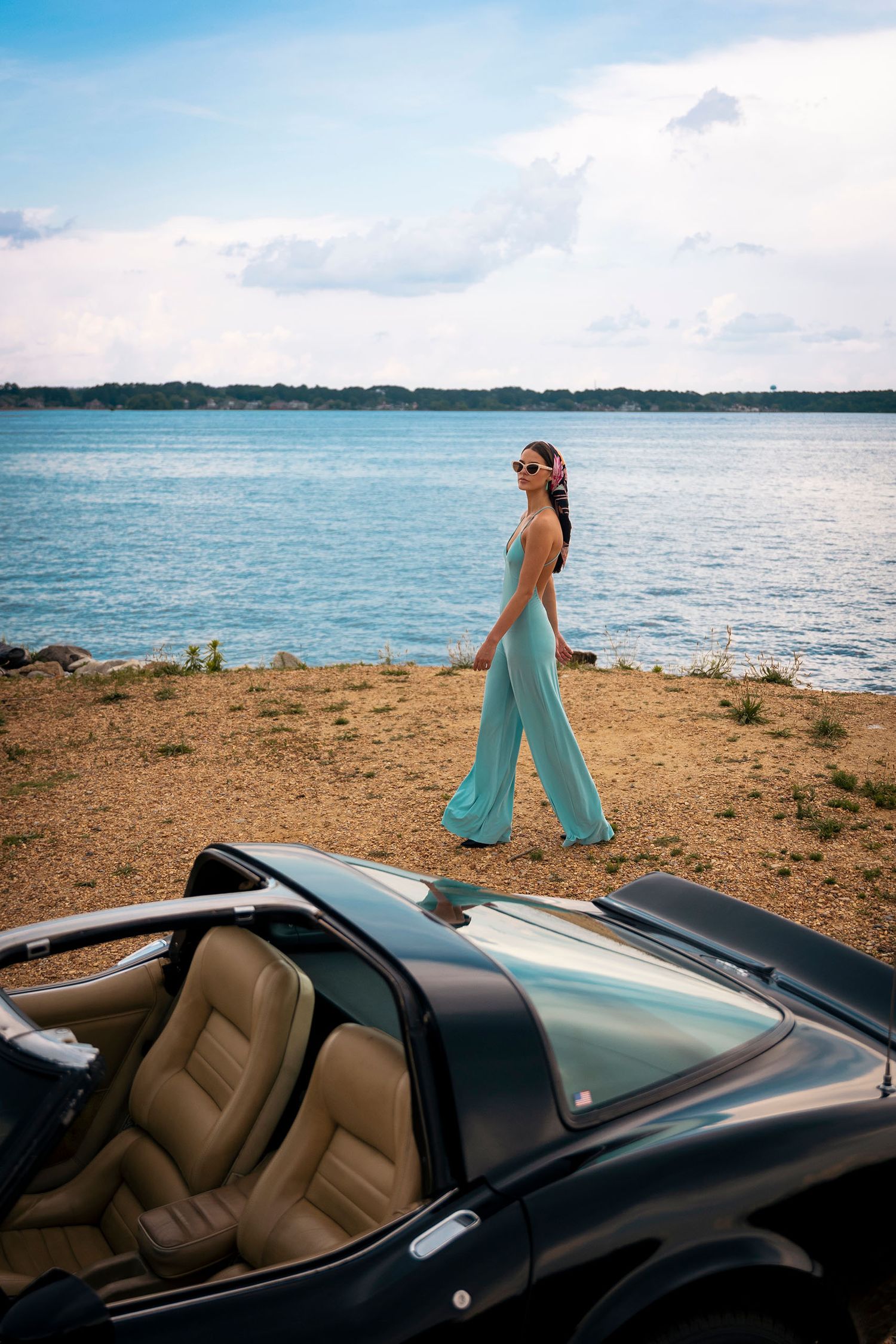Elegant woman in teal wide-leg jumpsuit walking near lakeside with vintage convertible car.