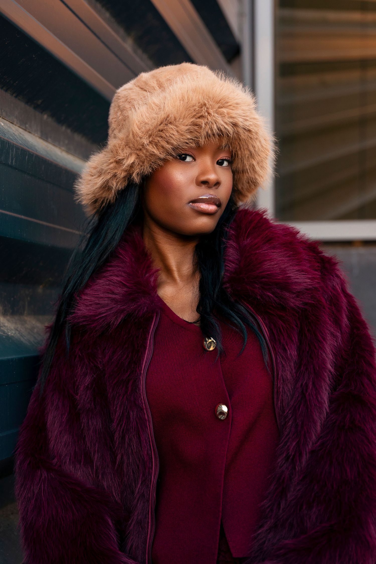 Woman in burgundy fur coat and tan fur hat poses against dark textured wall.