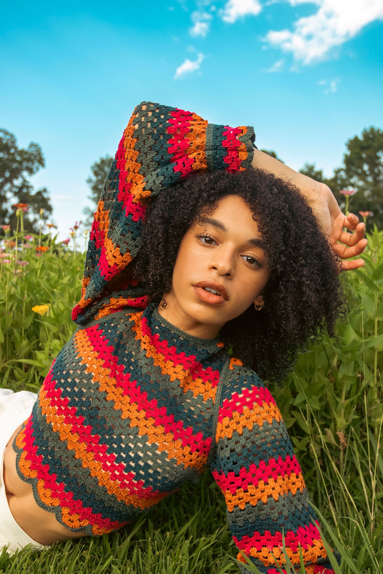 Woman with natural afro hair wearing multicolor crochet hoodie sitting in a wildflower meadow.