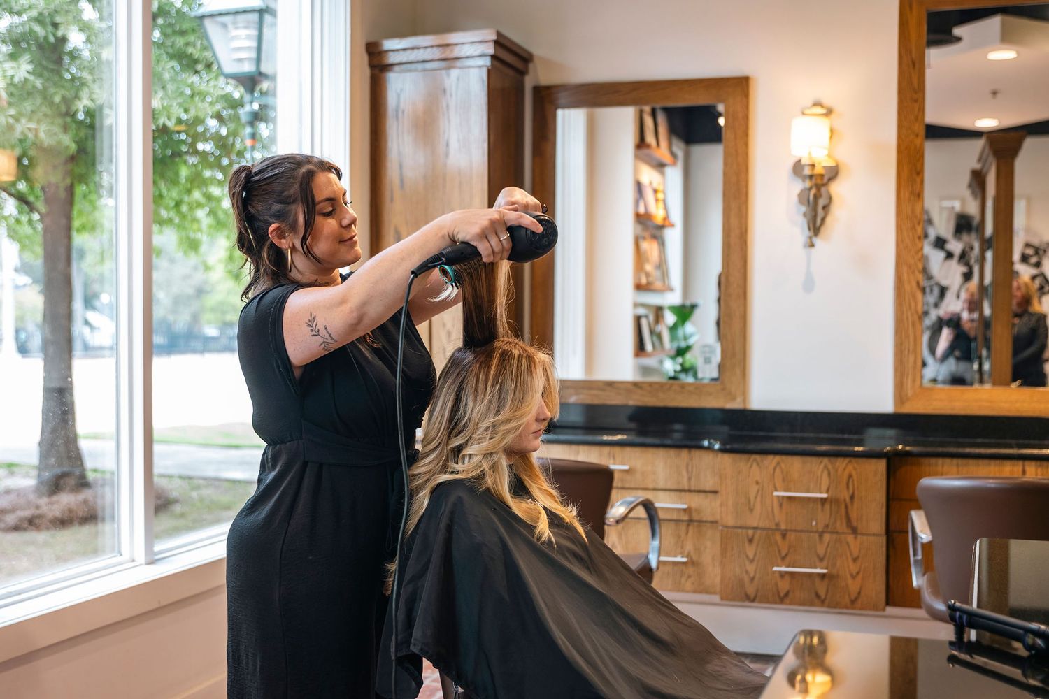 Hair stylist blow-drying a blonde client's hair in a modern salon near a large window.