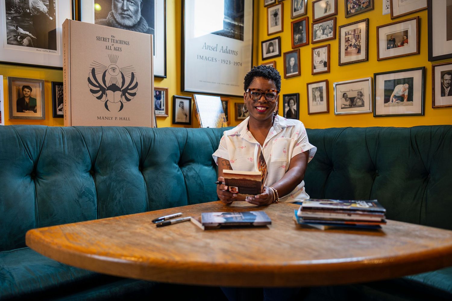 A smiling woman sits at a wooden table in a cozy cafe surrounded by books and framed photos.