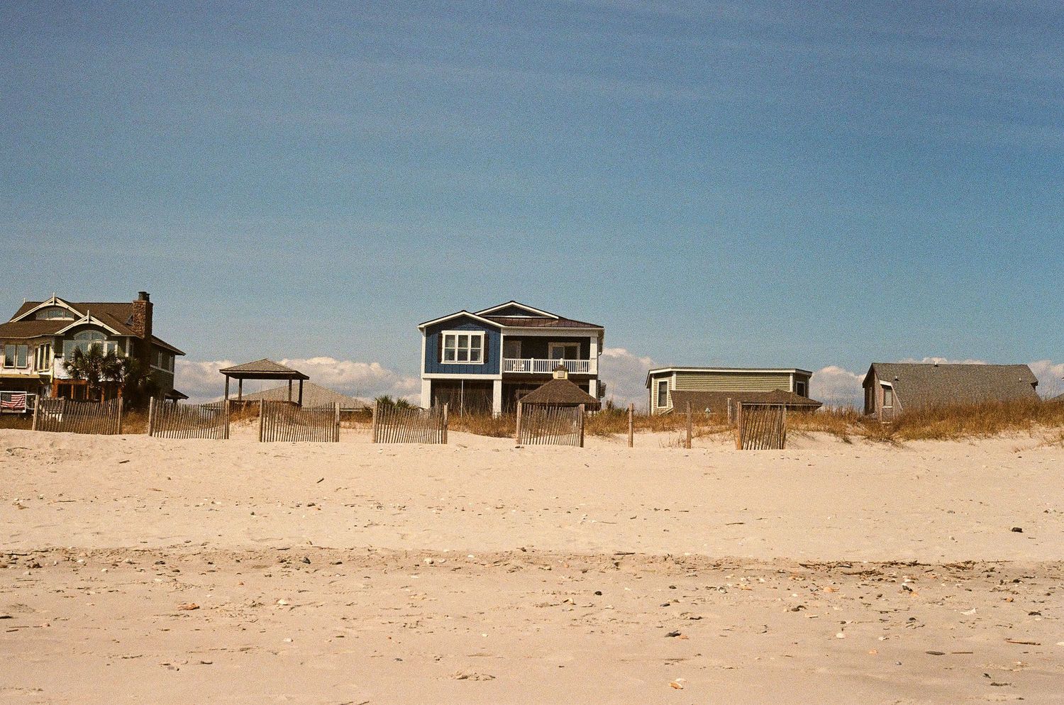 Colorful beach houses stand behind sandy dunes under a bright blue sky at the shore.