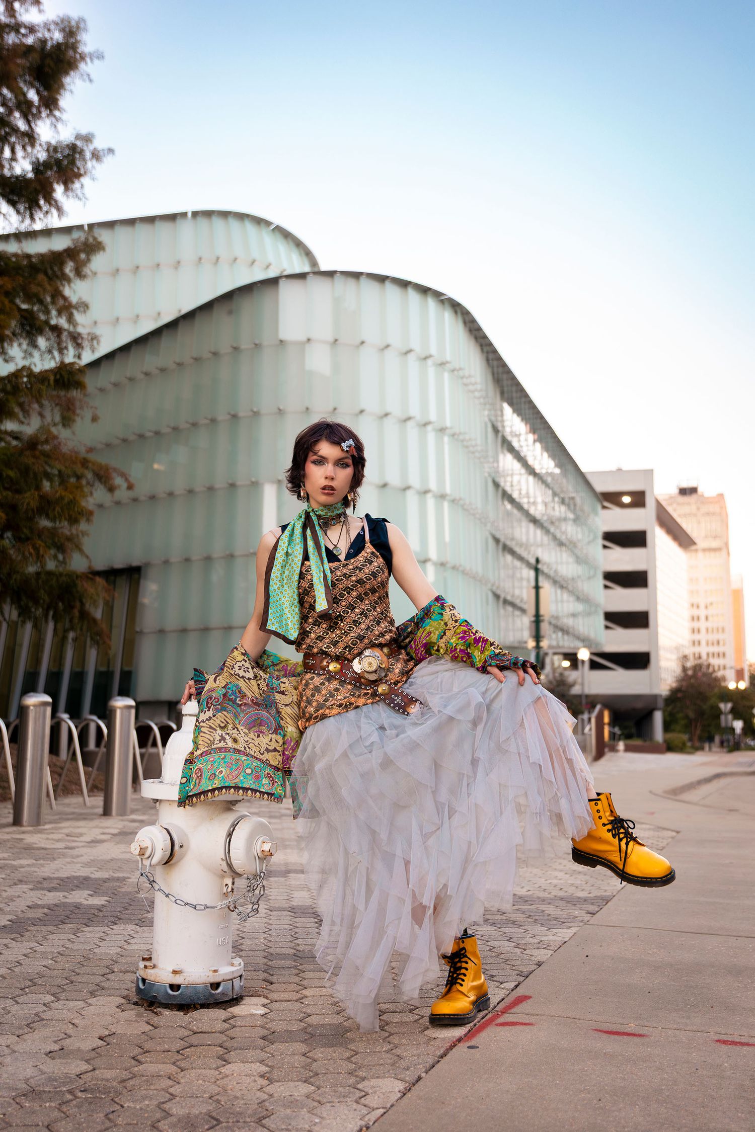 Model in bohemian layered outfit with yellow boots sitting on fire hydrant near modern building.