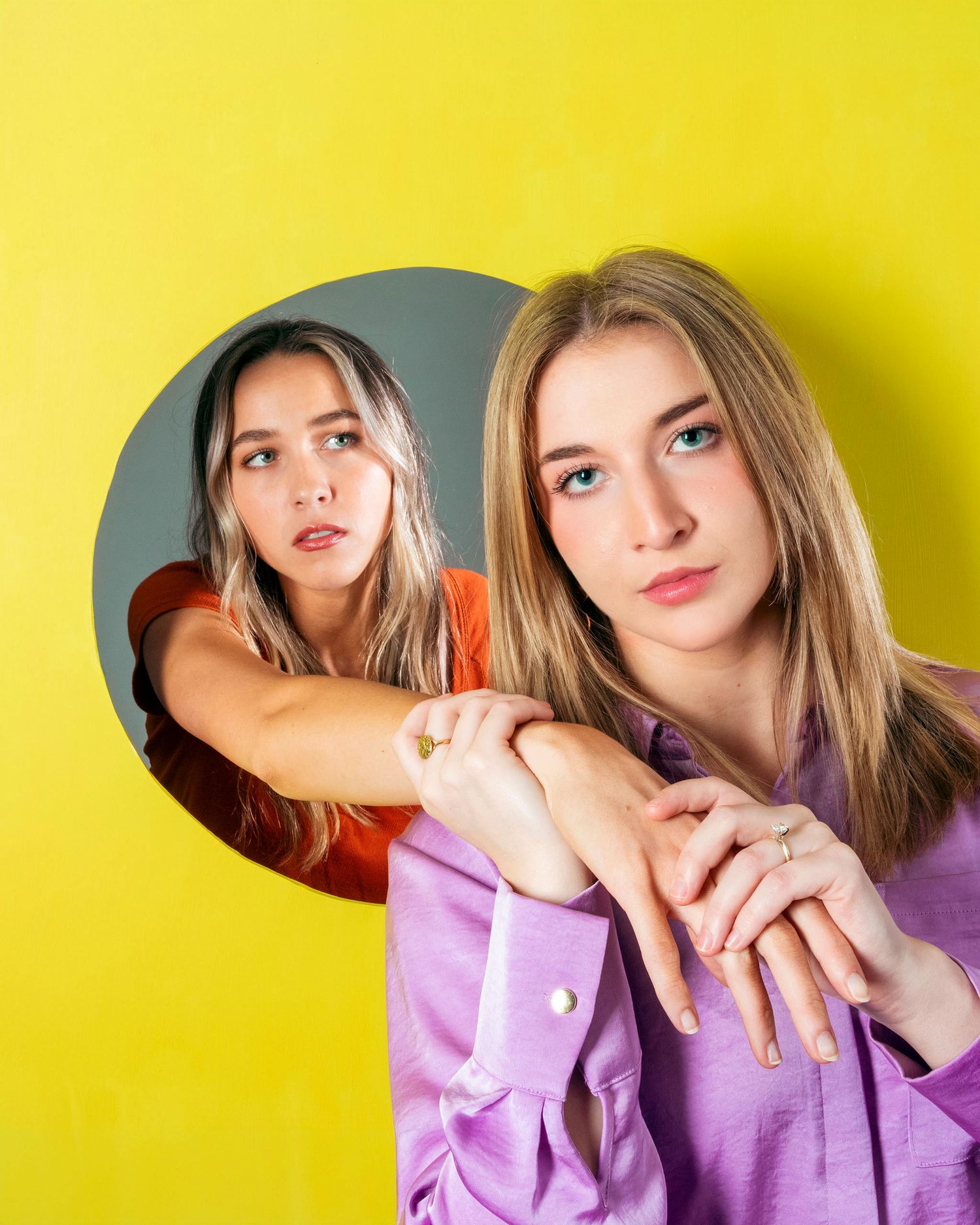 Two blonde women pose with circular wall cutout against bold yellow background.
