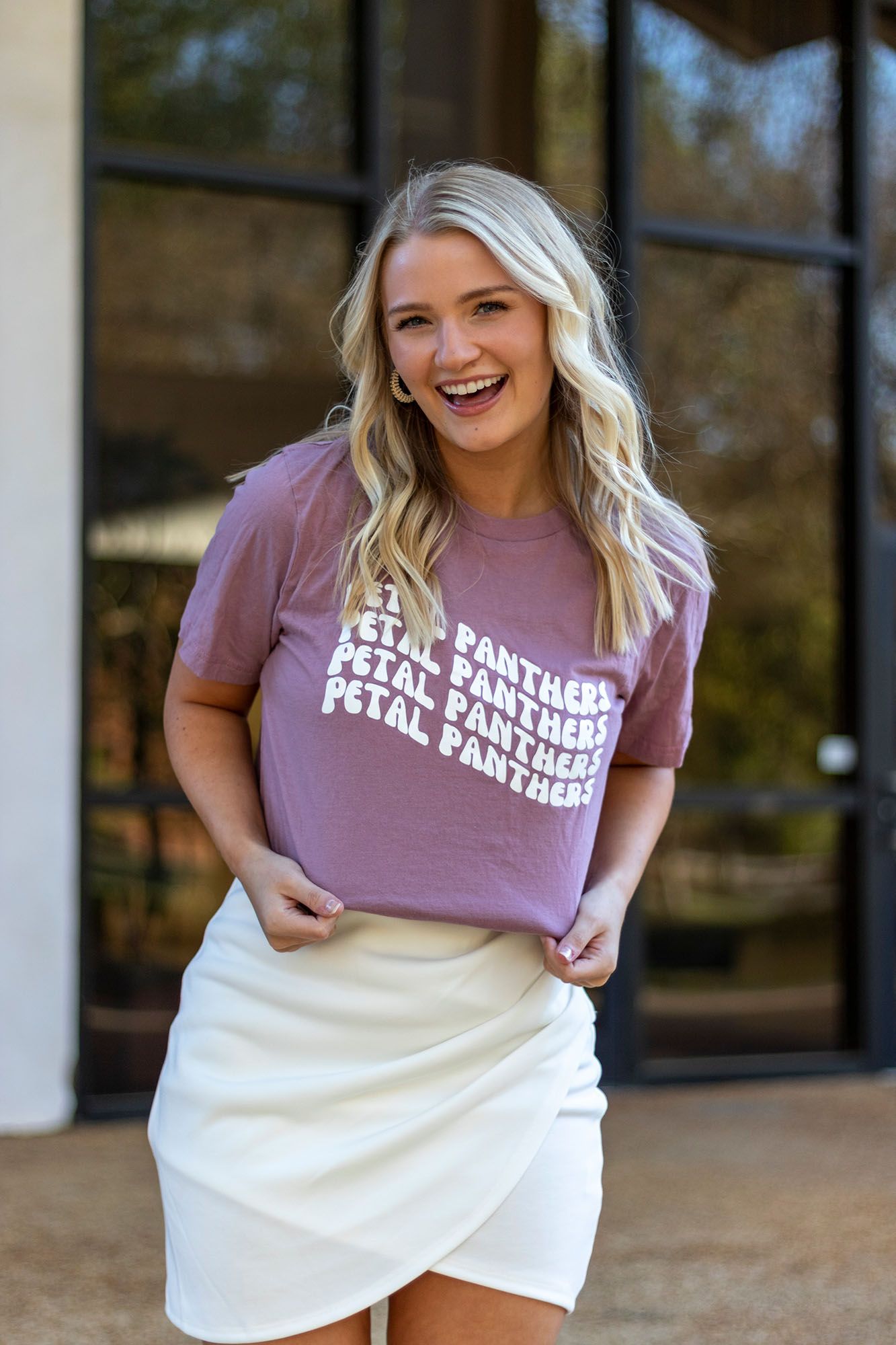 Laughing blonde woman in mauve Petal Panthers graphic tee and white wrap skirt poses outdoors.