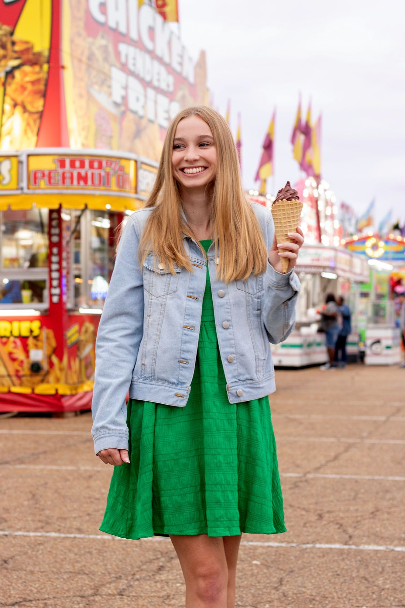 Smiling blonde girl in green dress and denim jacket holding ice cream cone at carnival.
