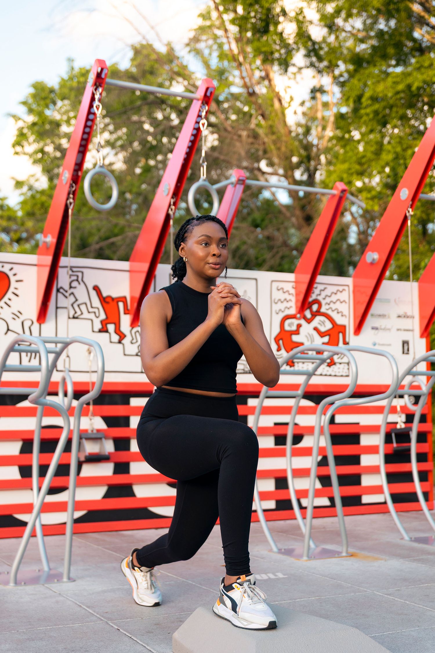 A Black woman in black athletic wear performs a lunge at an outdoor gym with red equipment.