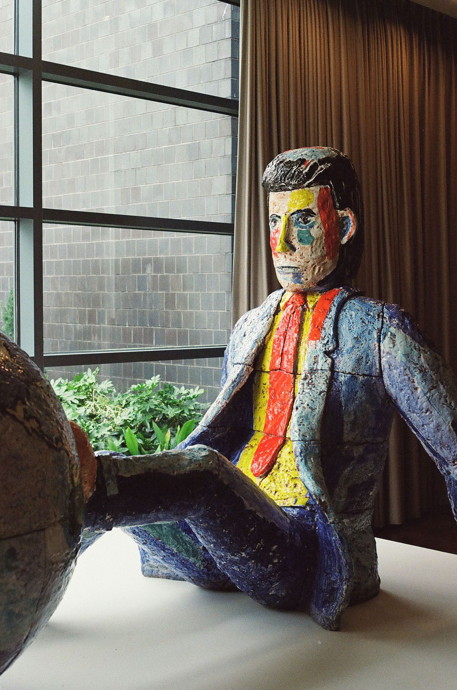Colorful painted ceramic sculpture of a seated man with vibrant red tie and yellow shirt displayed indoors.