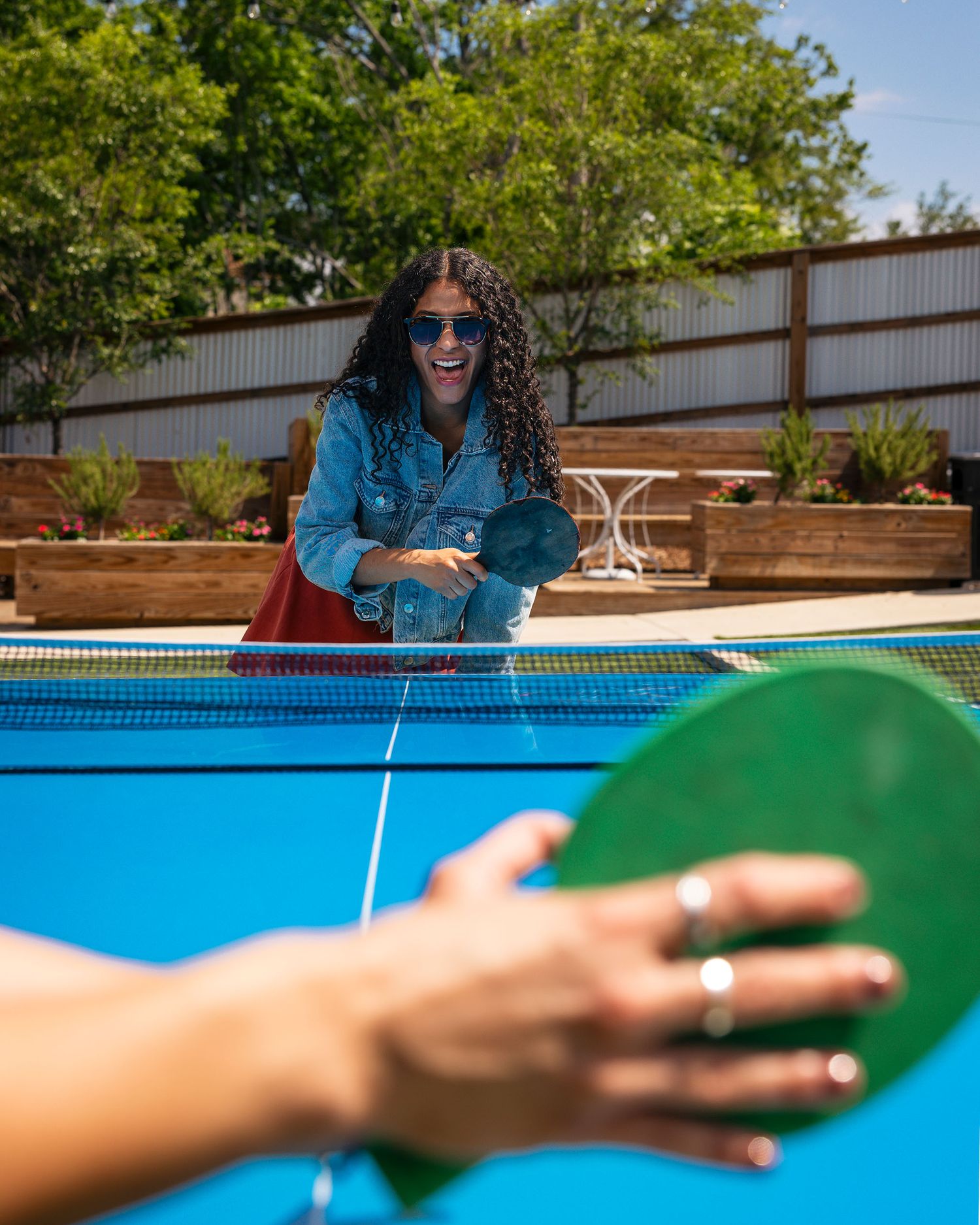 A laughing woman in a denim jacket plays ping pong outdoors on a sunny day.
