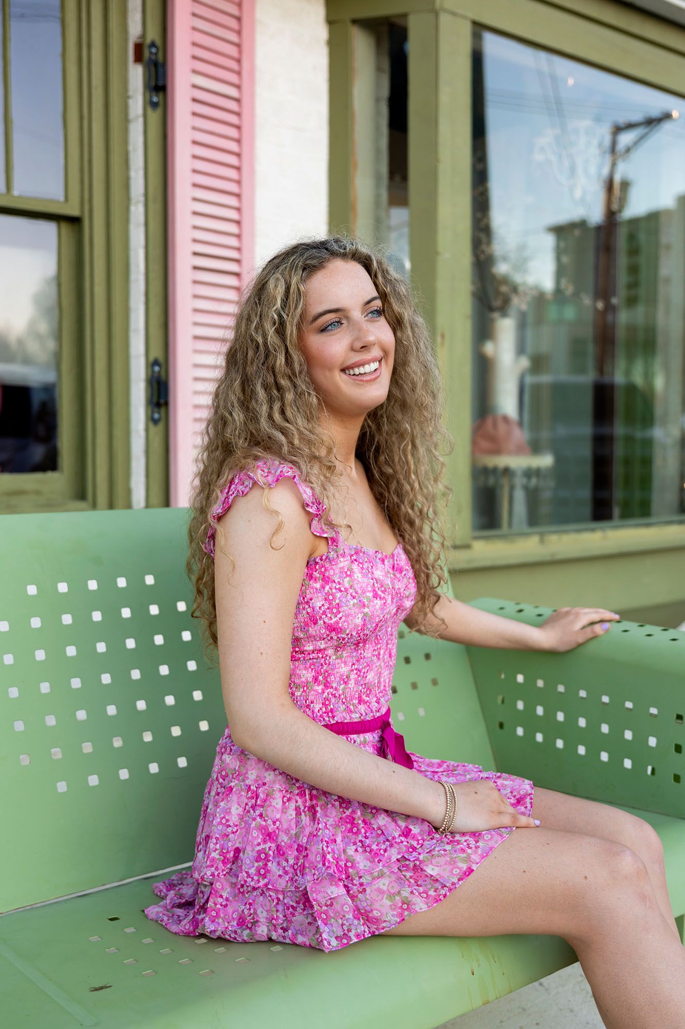 Cheerful young woman in pink floral dress sits on a green metal bench outside a shop.