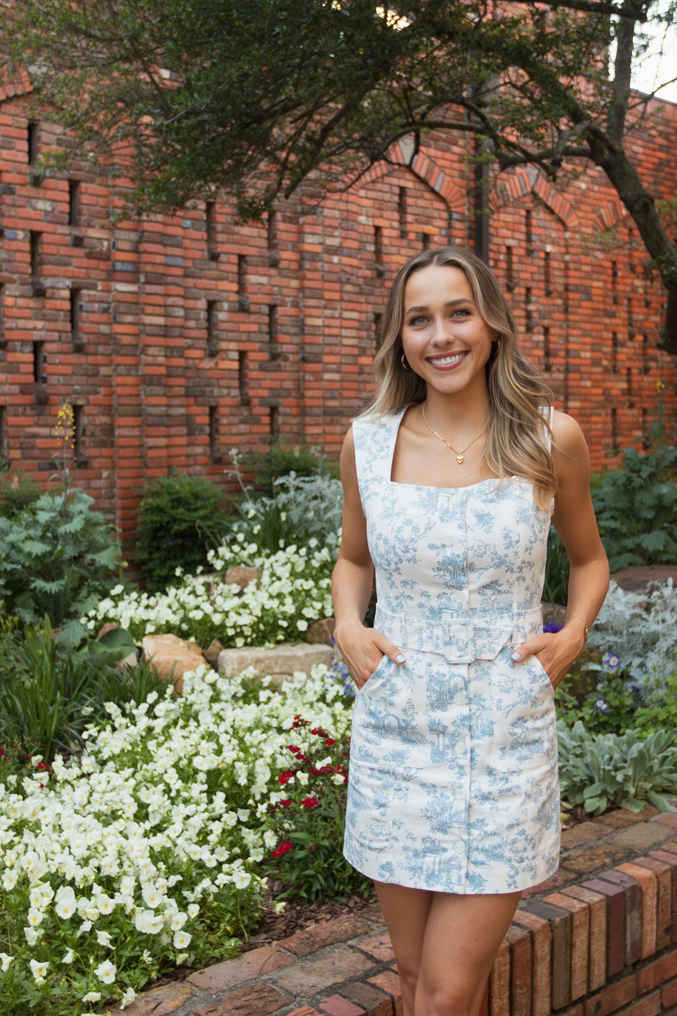 Smiling blonde female graduate in blue floral dress stands confidently before brick garden wall.
