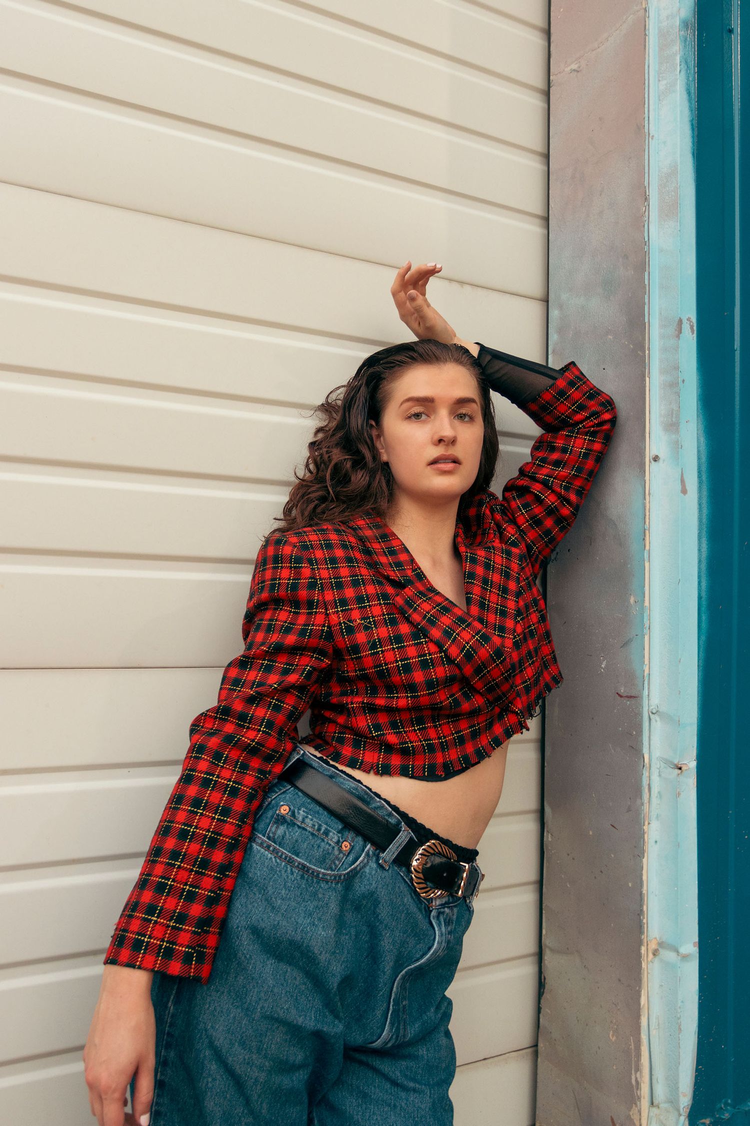 Woman in red plaid crop top and wide-leg jeans leans against a white corrugated wall.