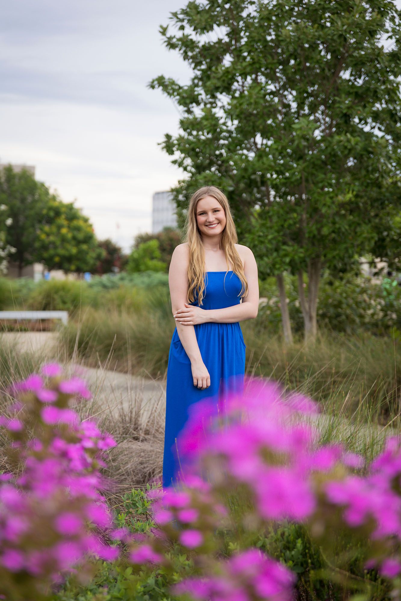 Young woman in a strapless royal blue gown stands among pink wildflowers in a garden.