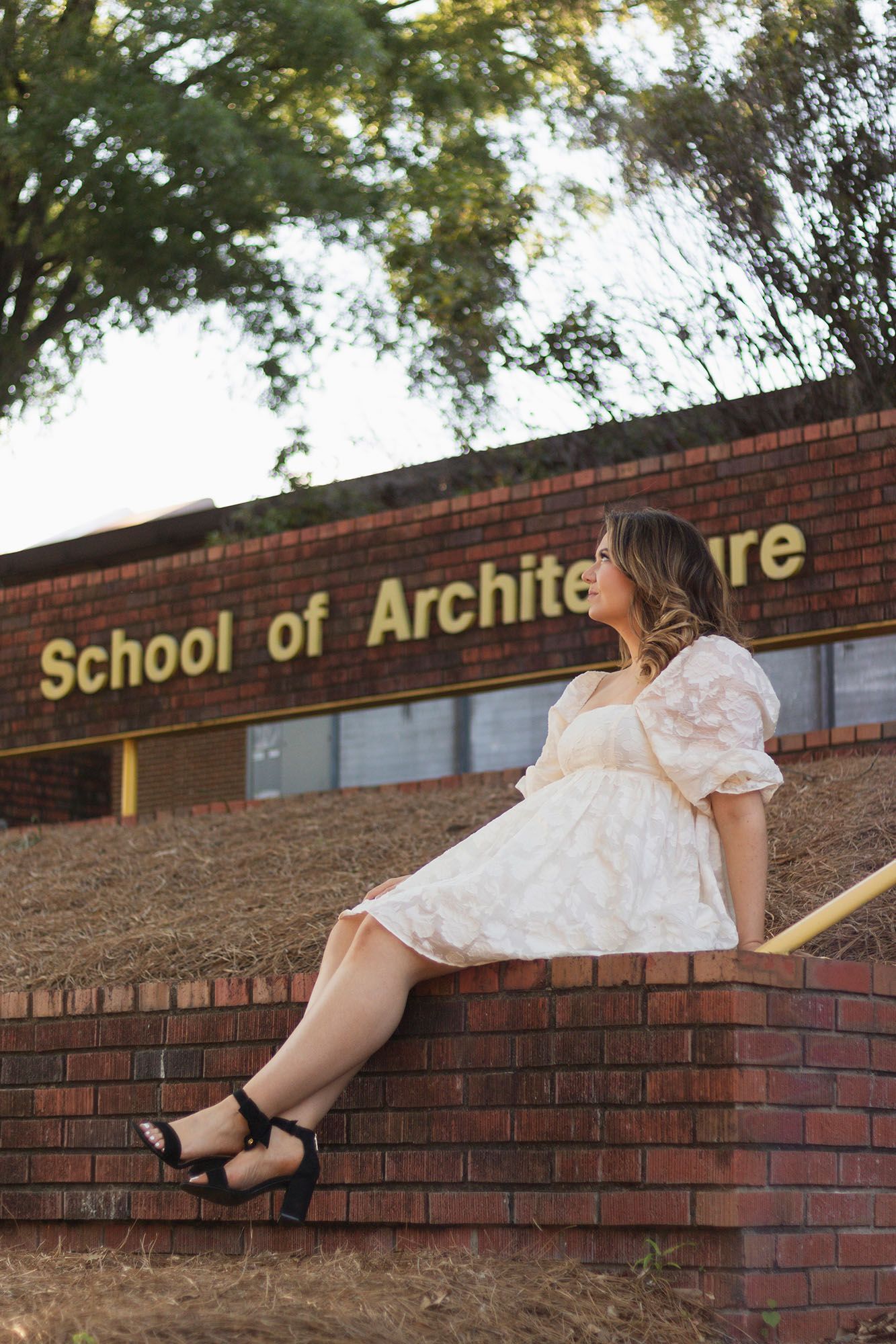 Graduate in white dress sits on brick wall outside School of Architecture building.