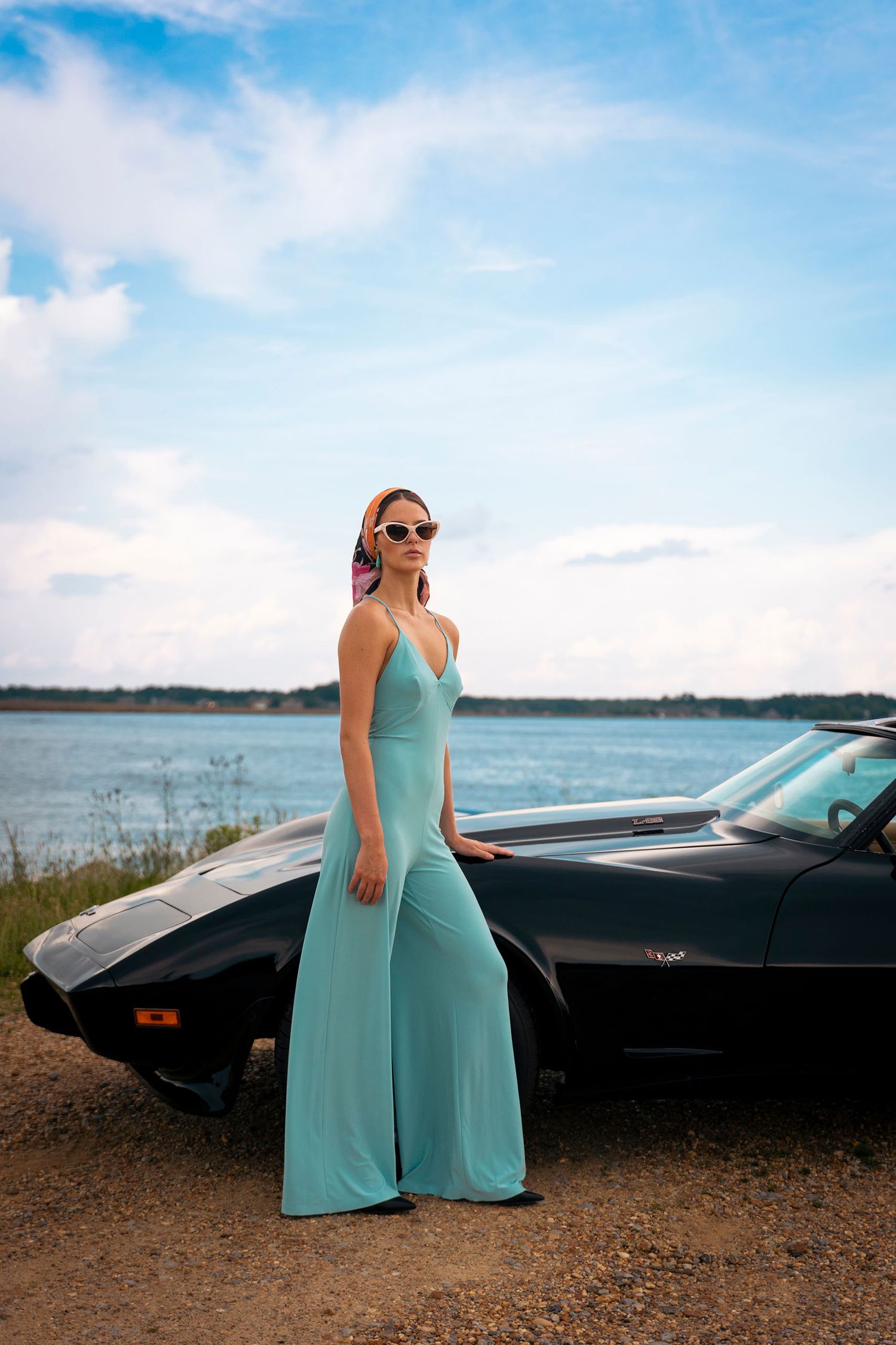 Woman in mint green wide-leg jumpsuit posing beside classic black convertible near waterfront.