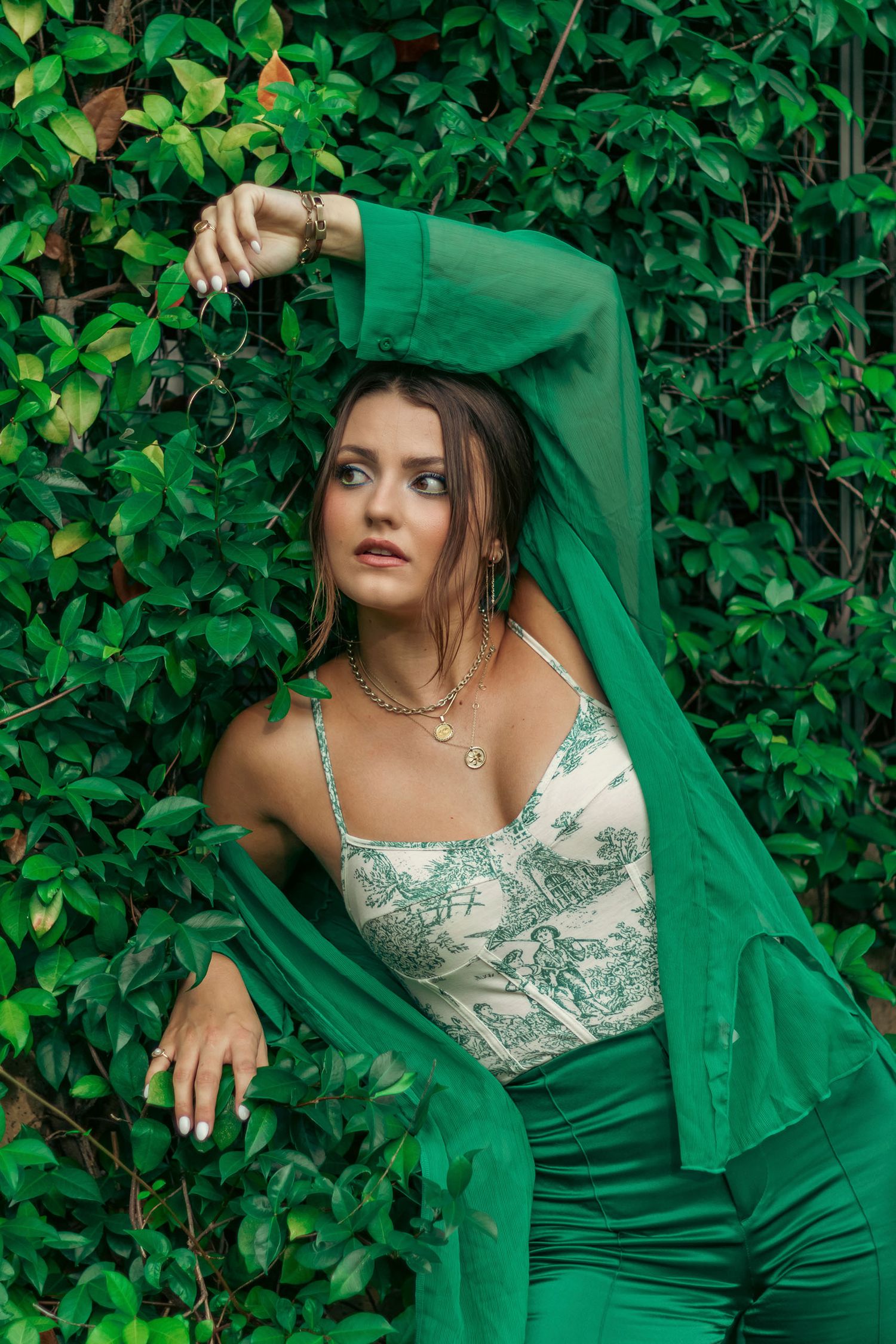 Woman in green outfit and white floral bodysuit poses against lush ivy-covered wall.