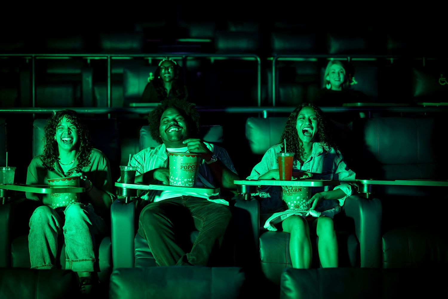 Group of friends laughing and screaming while watching a movie in a theater under green lighting.