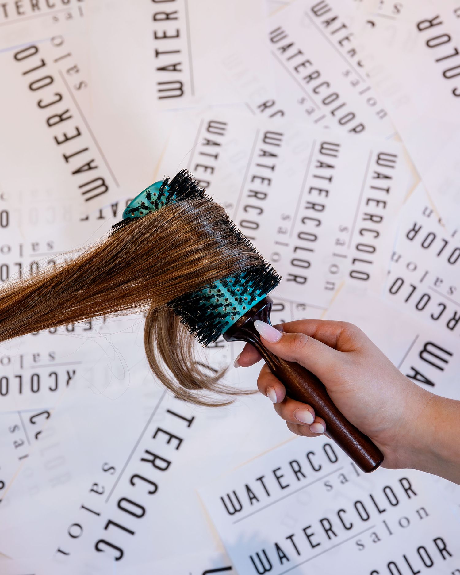 Hand holding a teal round brush styling brown hair over Watercolor Salon branded paper background.
