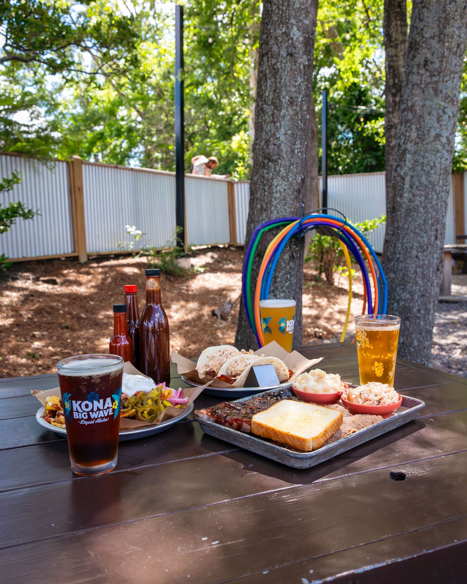 An outdoor picnic table with Kona Big Wave beers, BBQ food, and a colorful rainbow decoration.