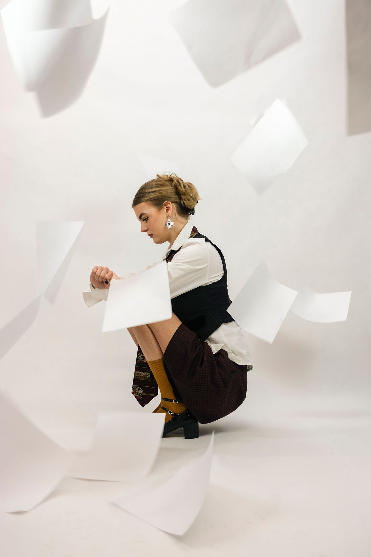 Woman crouching amid flying white papers in a dynamic editorial fashion photo with white background.