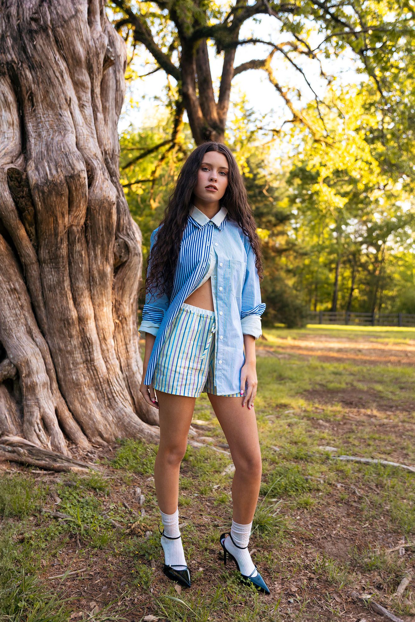 Young woman in striped blue outfit posing beside large tree trunk in park setting.