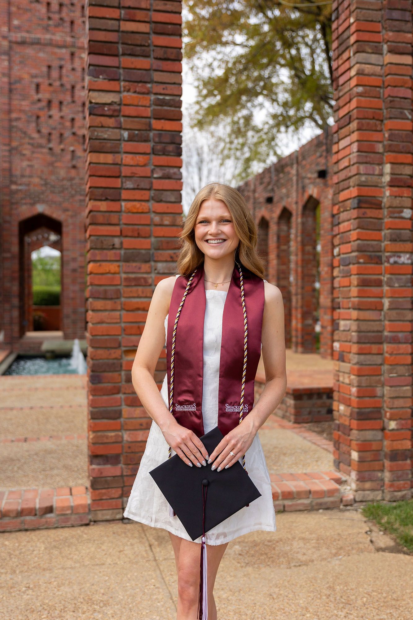 Smiling female graduate in white dress with maroon stole holds cap at brick university campus.
