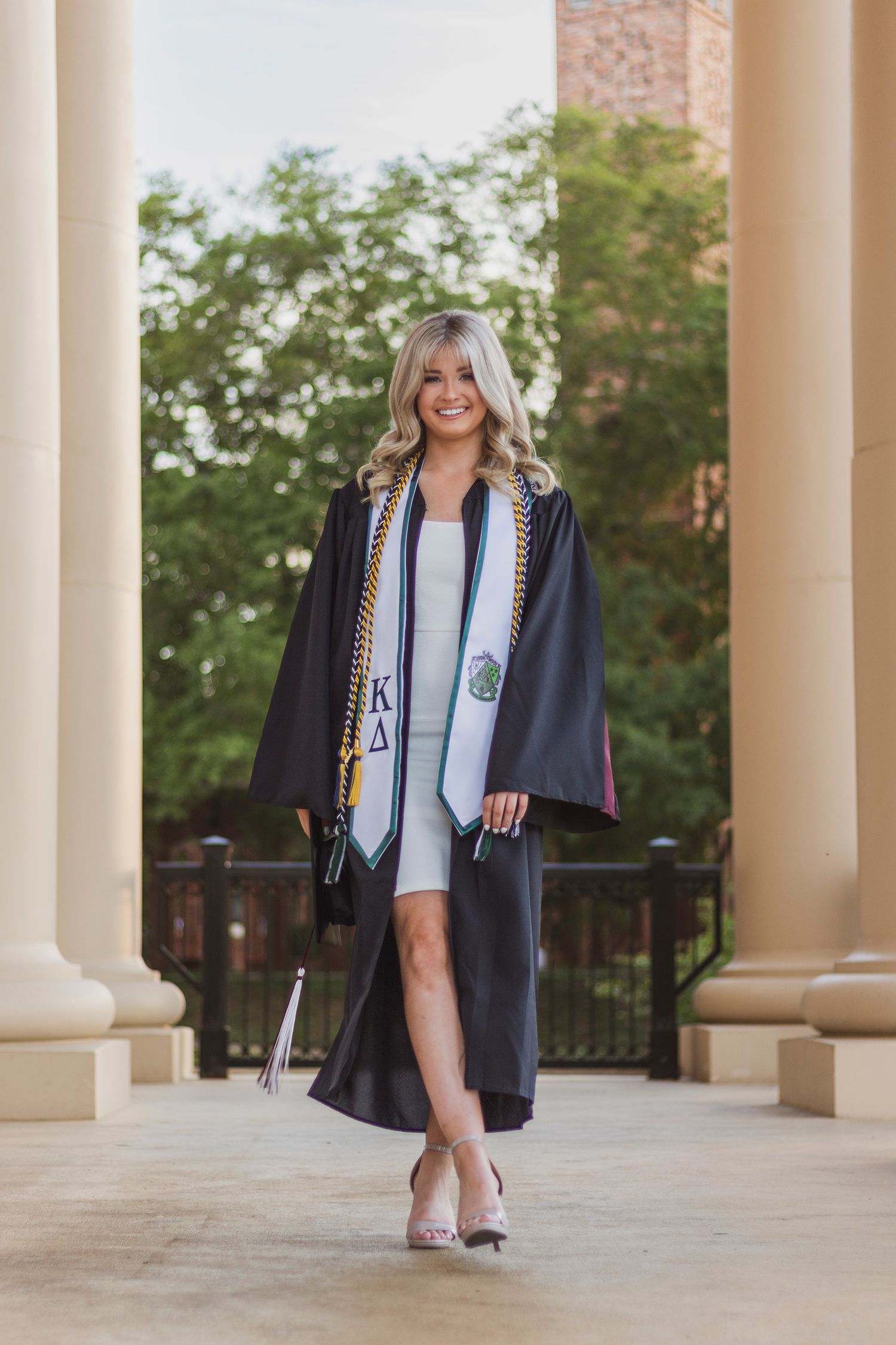 Smiling blonde graduate walks in black gown with Kappa Delta stoles between white campus columns.