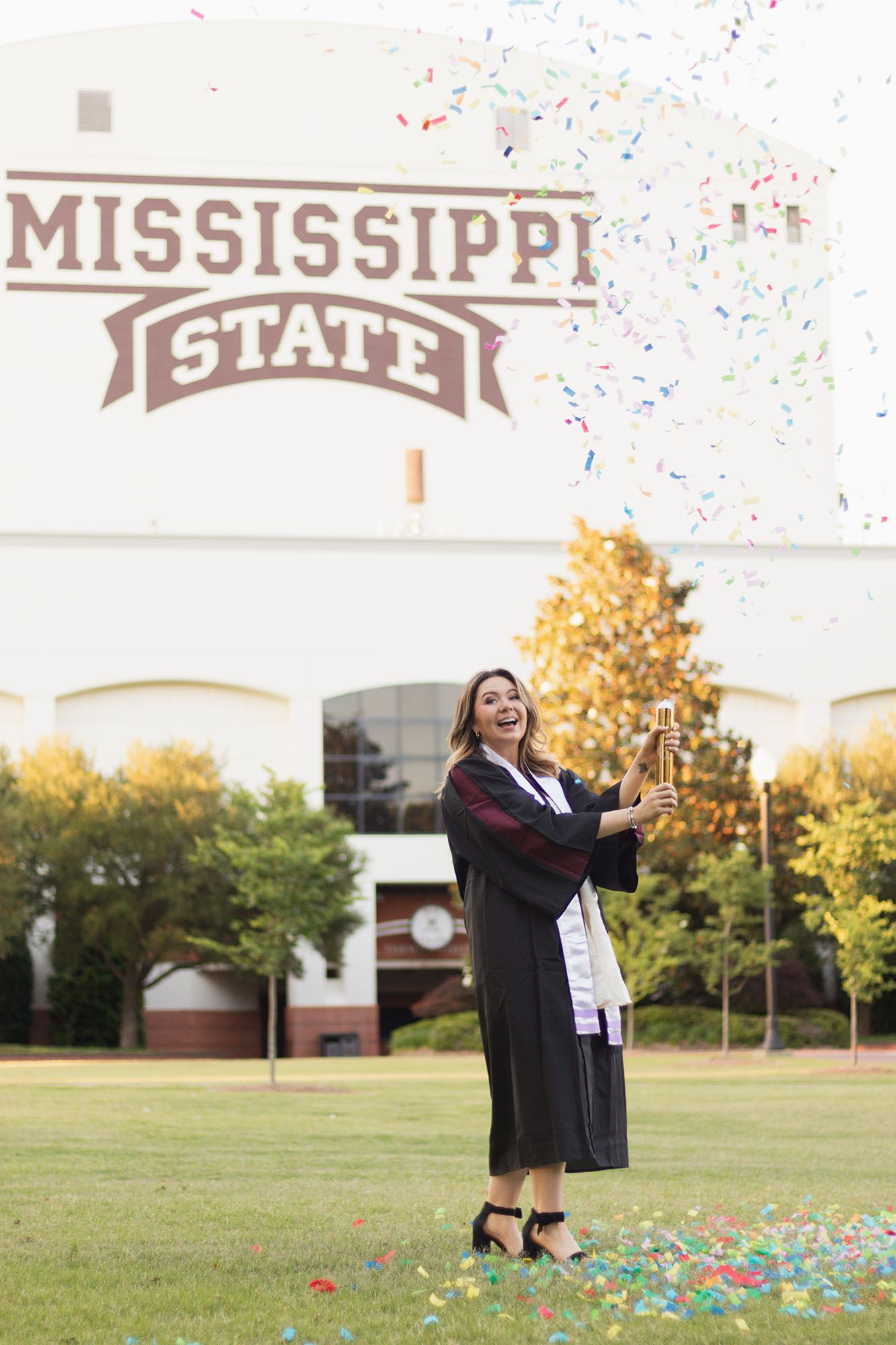 Graduate in cap and gown celebrates with confetti in front of Mississippi State sign.