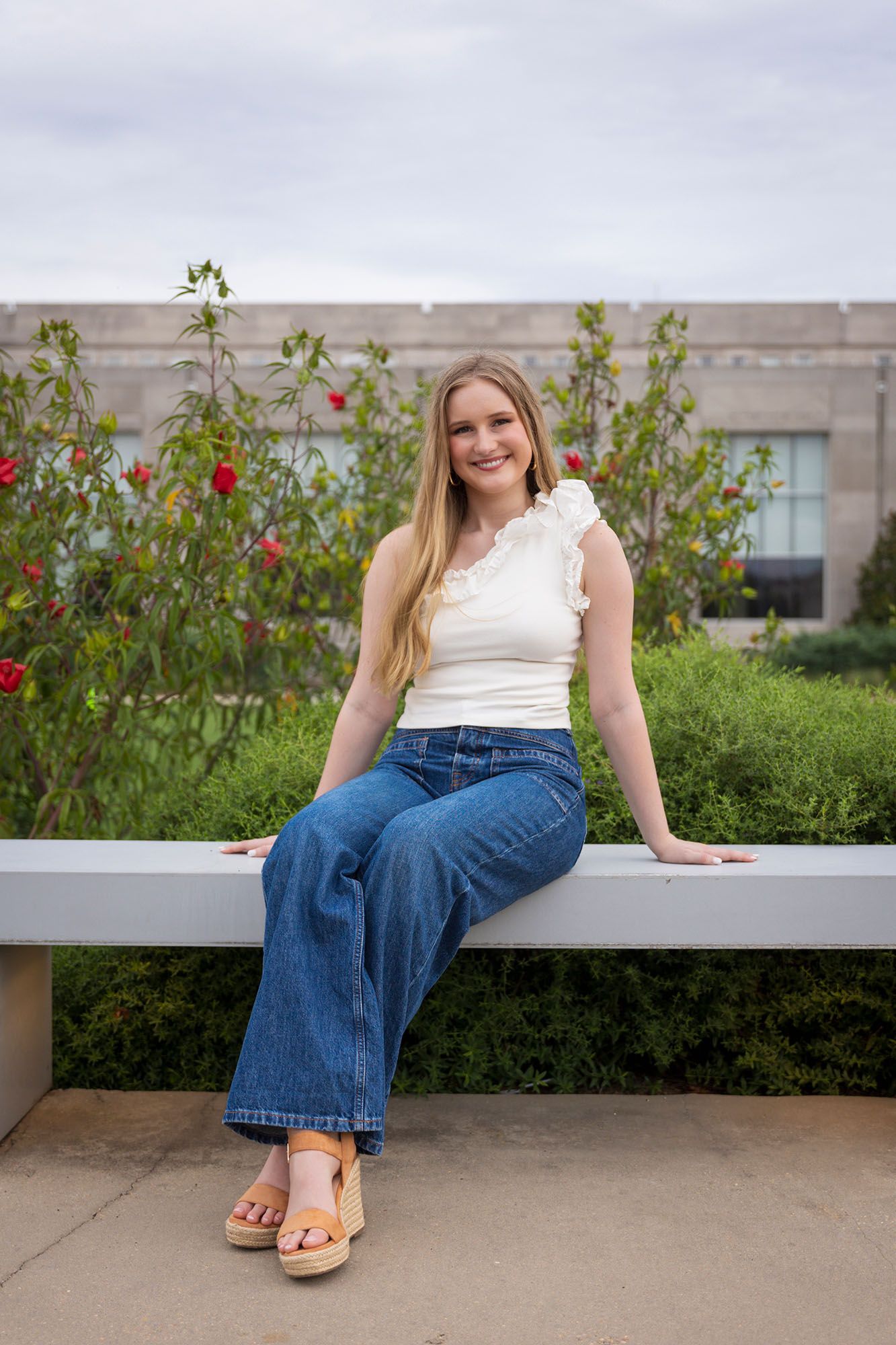 Young blonde woman in white top and wide-leg jeans sits on a bench in a rose garden.