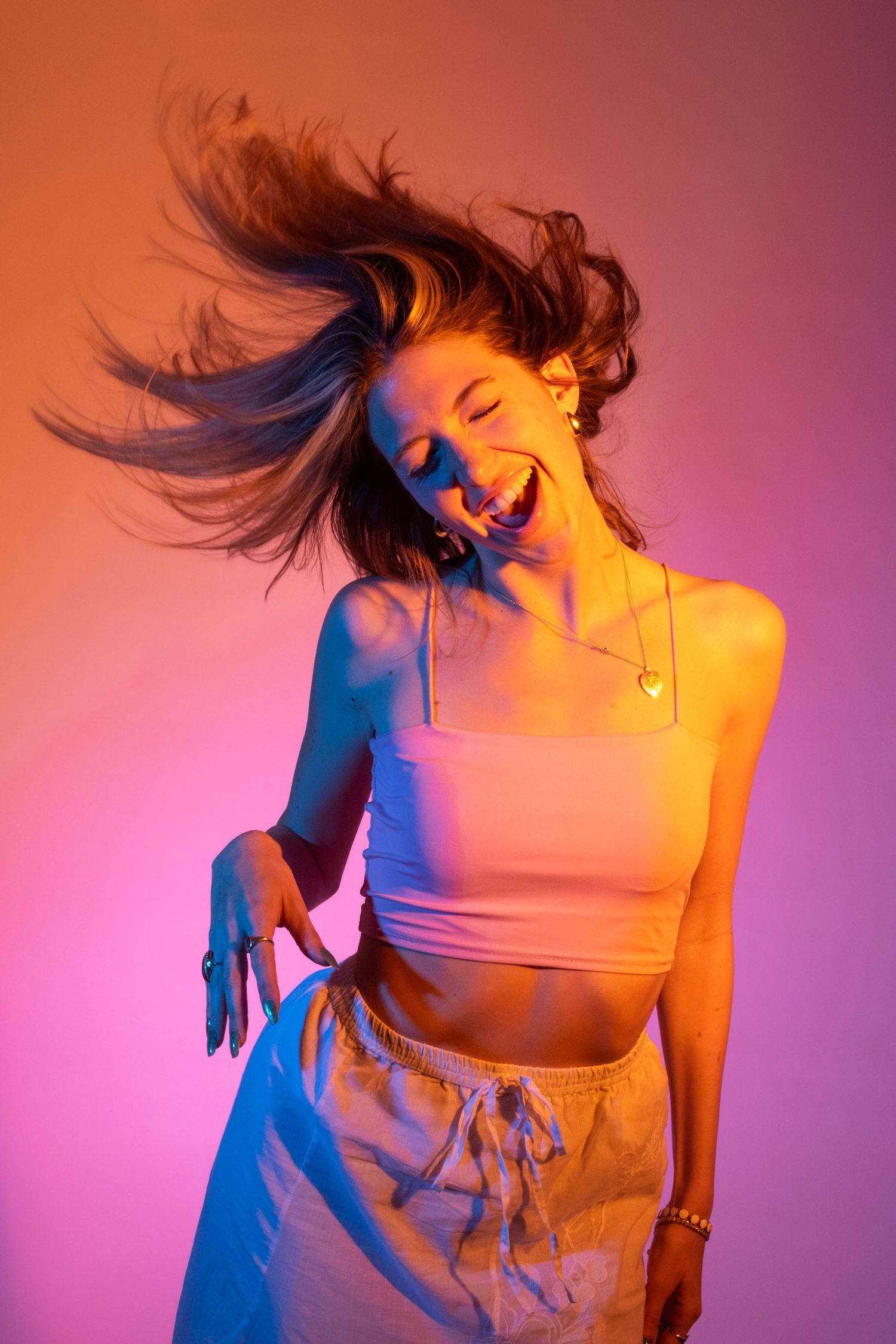 Young woman in white crop top laughs with hair flying in pink and purple neon light.
