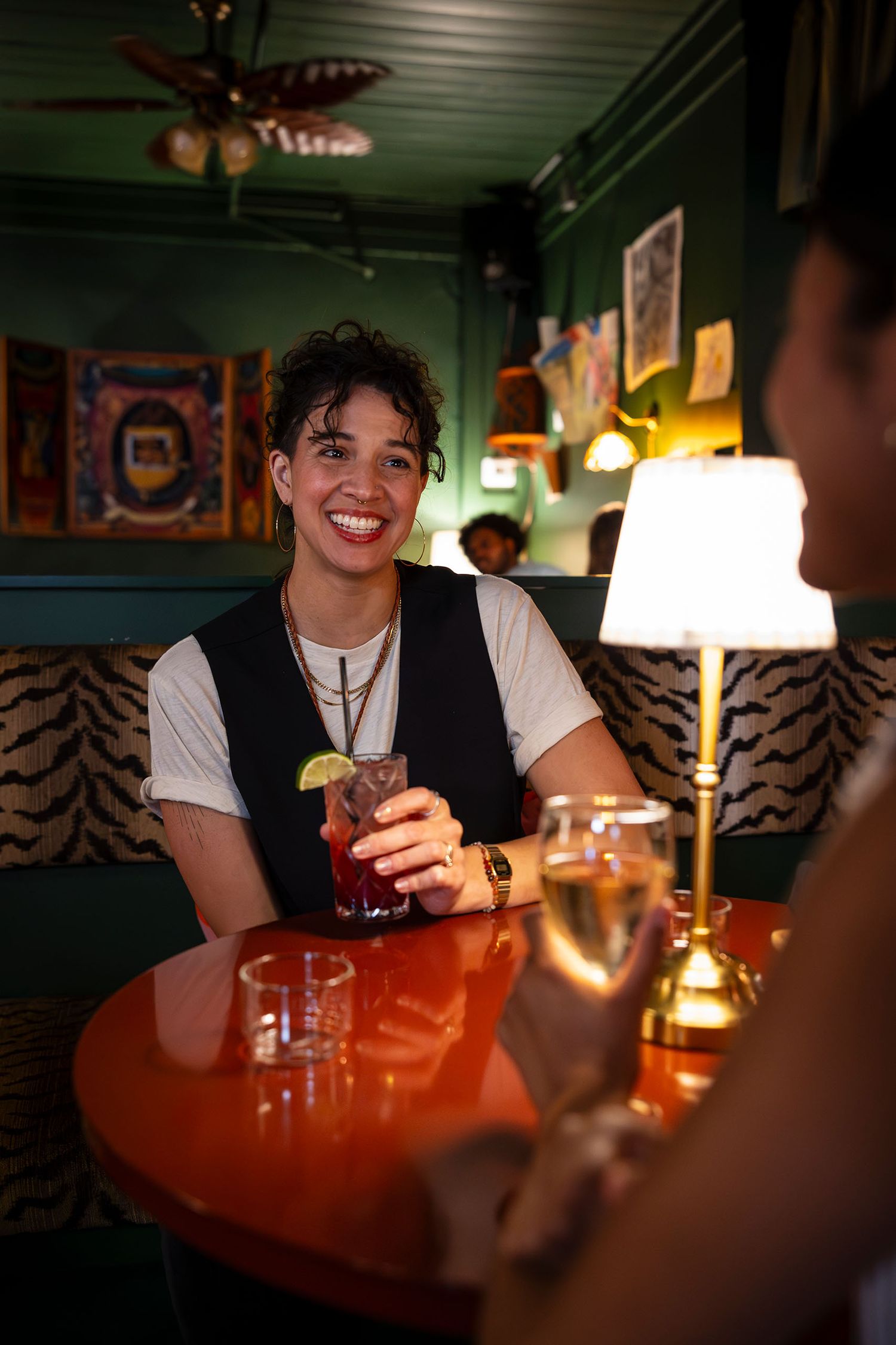 Smiling woman enjoying a cocktail in a cozy green-walled bar with warm lighting.