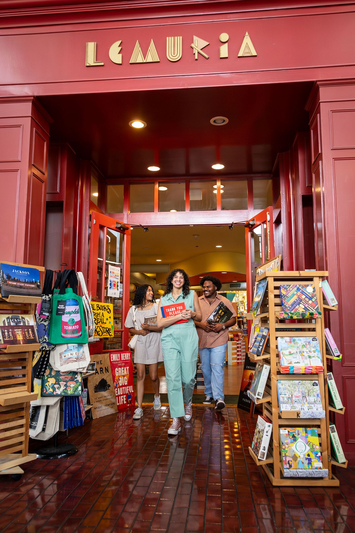 Three friends exit Lemuria bookstore carrying books through a vibrant red storefront entrance.