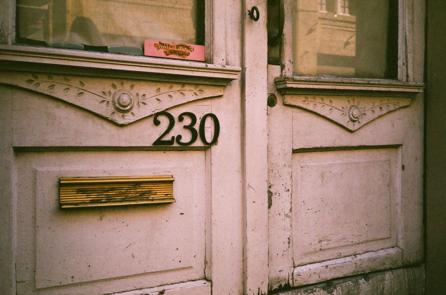 A weathered pink wooden door numbered 230 with ornate carvings and a brass mail slot.
