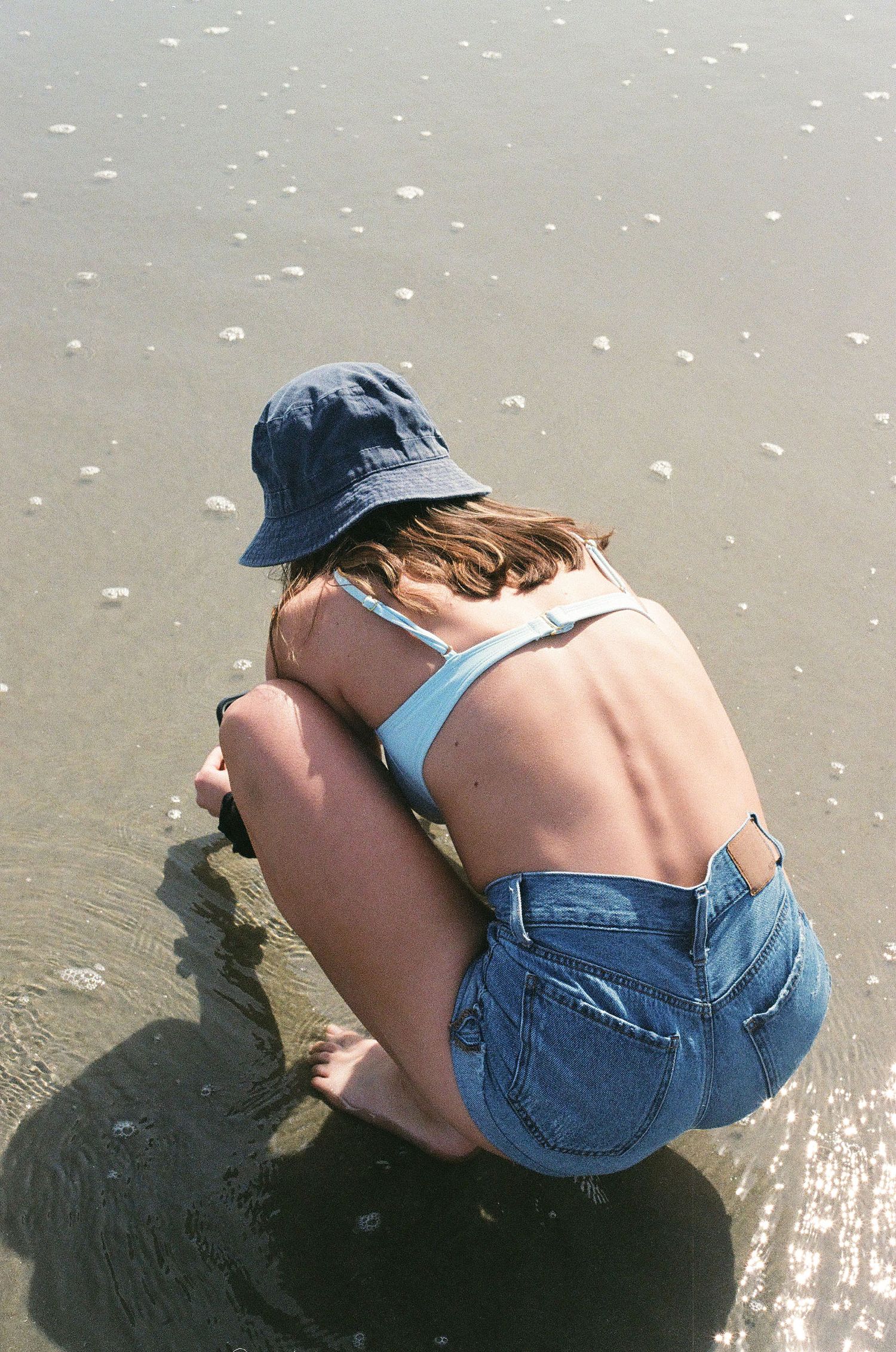 A woman in a blue bucket hat and denim shorts crouches on a wet sandy beach.