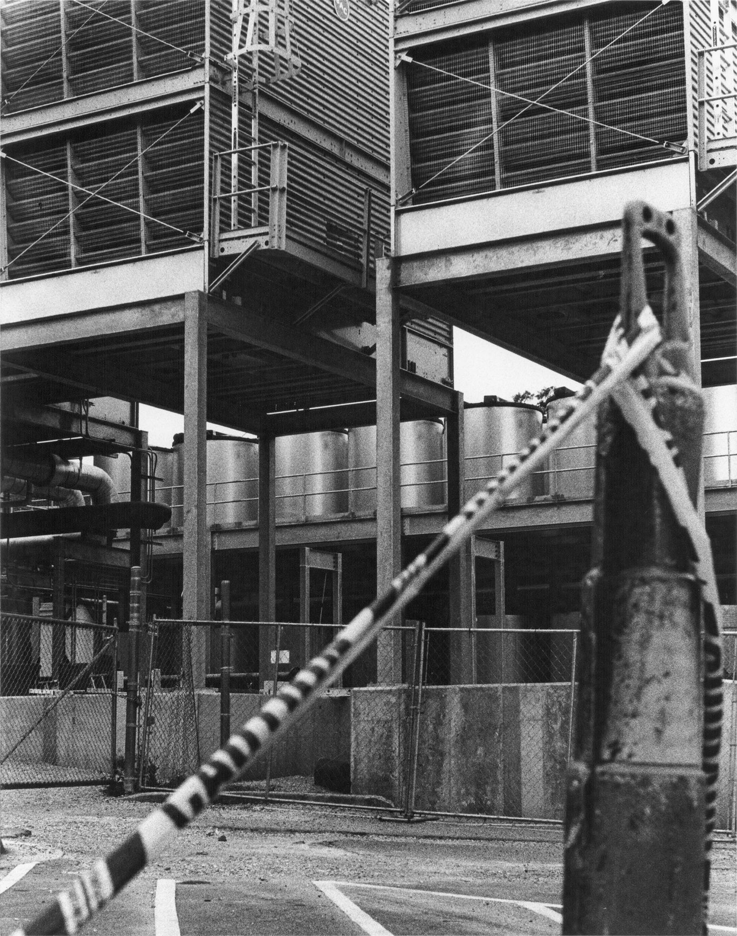 Black and white industrial facility exterior with large tanks, steel framework, and a chained entrance gate.